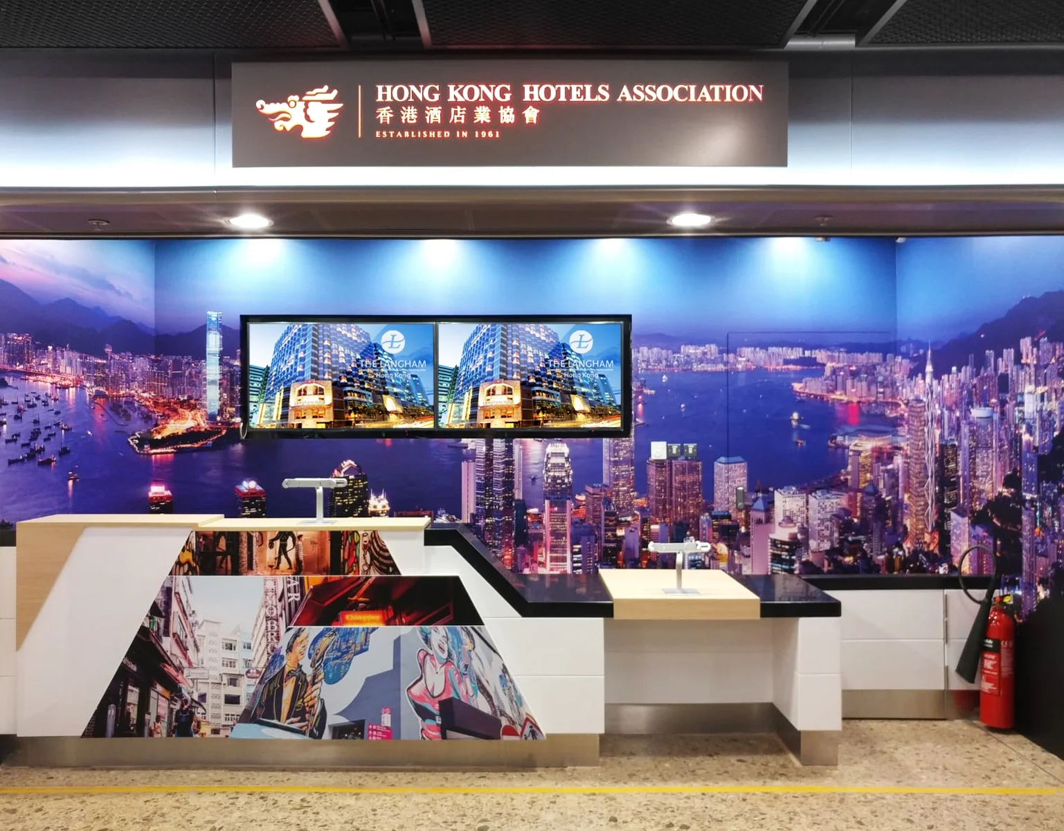 A Hong Kong Hotels Association booth with a vibrant cityscape mural background featuring a harbor and skyline. There are two monitors displaying a building in Hong Kong, and a counter with artwork depicting urban scenes and graffiti.