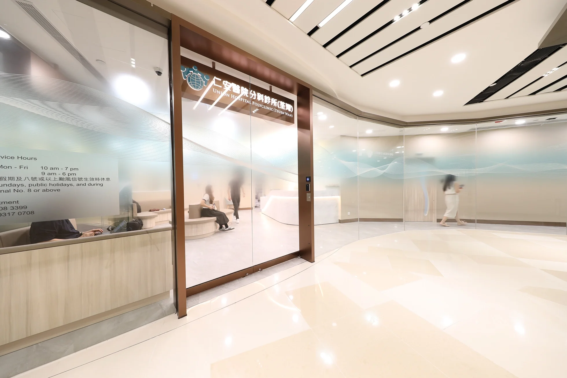 Modern hospital lobby with transparent glass walls, wooden accents, and a reception area. People are seated and walking, with overhead lighting and ceiling vents visible.