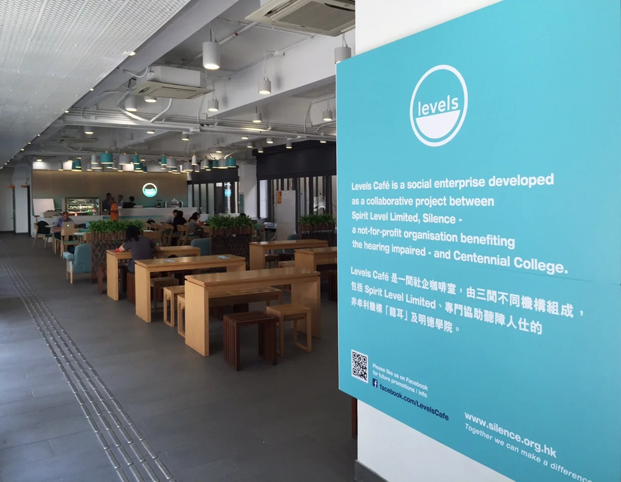 Interior of a modern café with wooden tables and chairs, some people seated, and a large blue informational sign on the right side explaining the café's social enterprise background with text in English and Chinese.