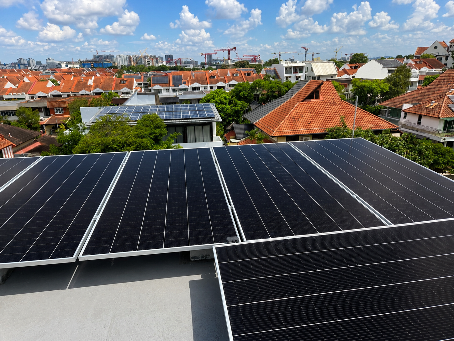 View of solar panels installed on rooftops in a suburban neighborhood with red tiled roofs, green trees, and a city skyline with construction cranes in the background under a partly cloudy sky.