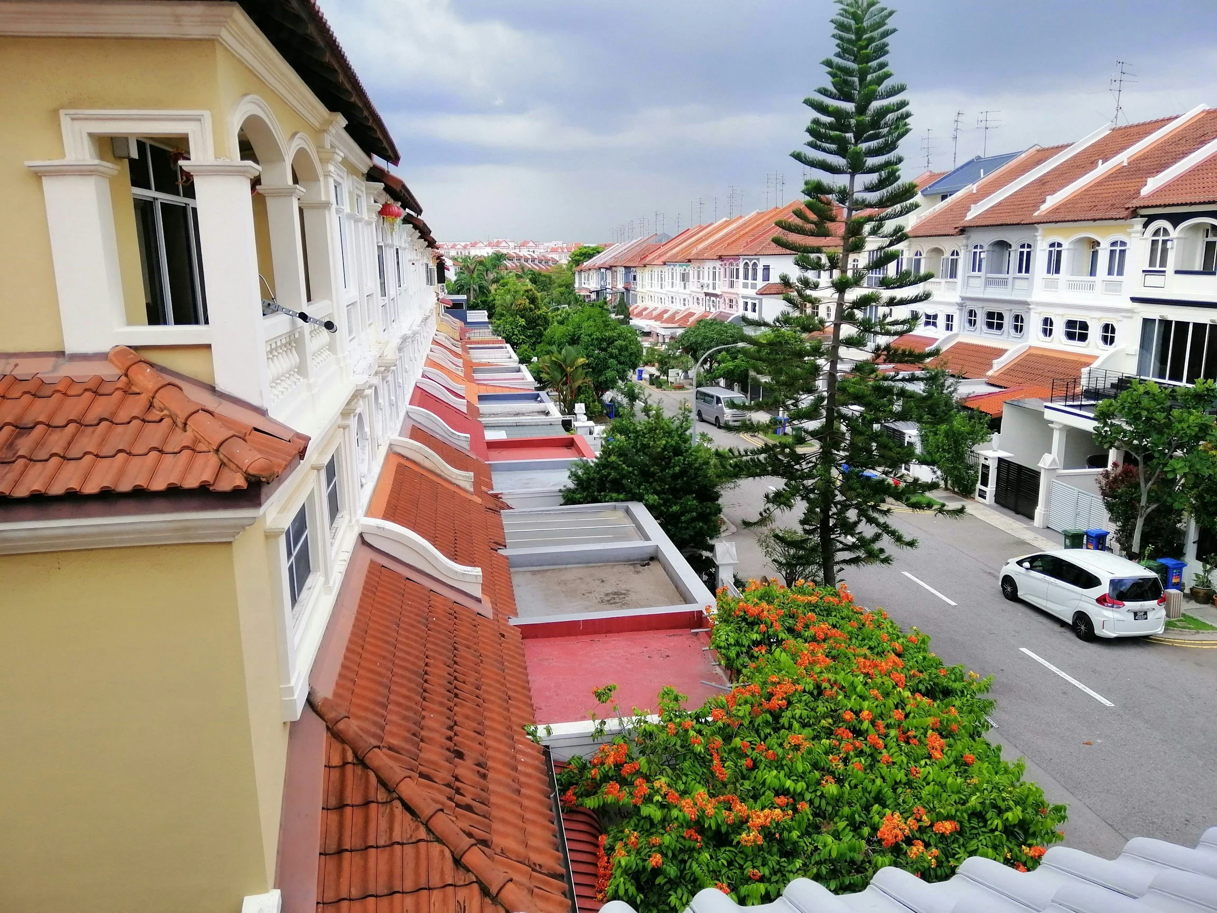 View of a residential neighborhood with multiple townhouses featuring white walls and orange-tiled roofs, a street with parked cars, lush green trees, and a cloudy sky overhead.