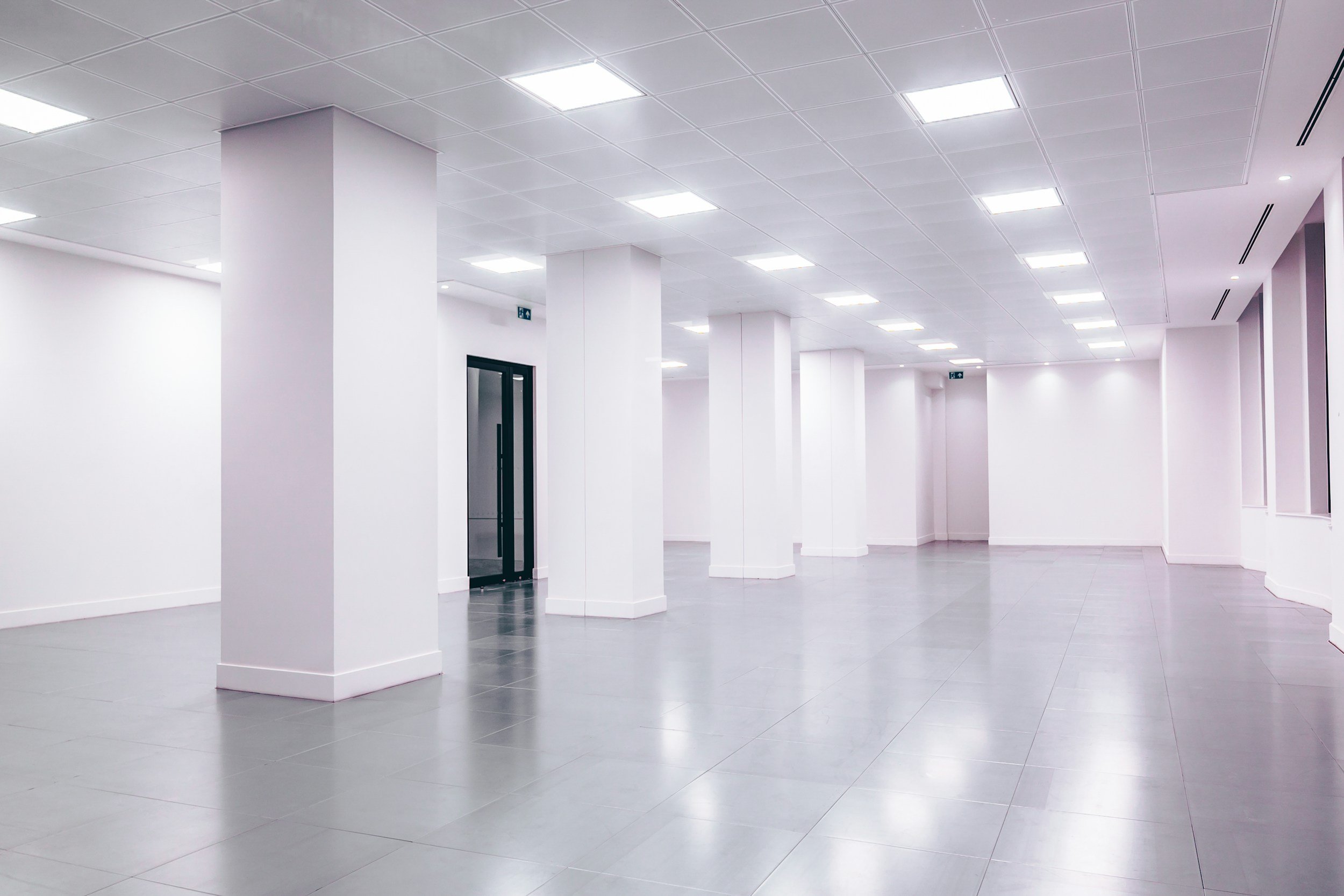 A spacious, modern, empty interior room with white walls, a tiled ceiling with evenly spaced square lights, and gray tiled flooring. There are white pillars and a black door in the background.