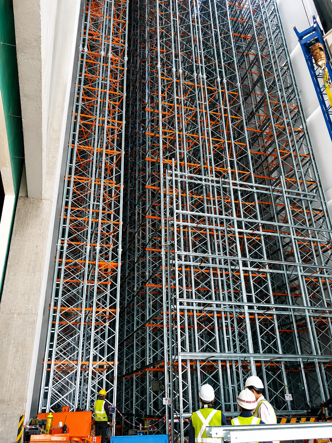 Construction workers in safety helmets and vests observing a large, complex steel storage rack system inside a building under construction.