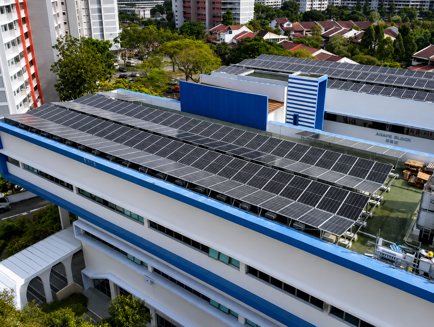 Aerial view of a building with a rooftop covered in solar panels, with trees and residential buildings in the background.