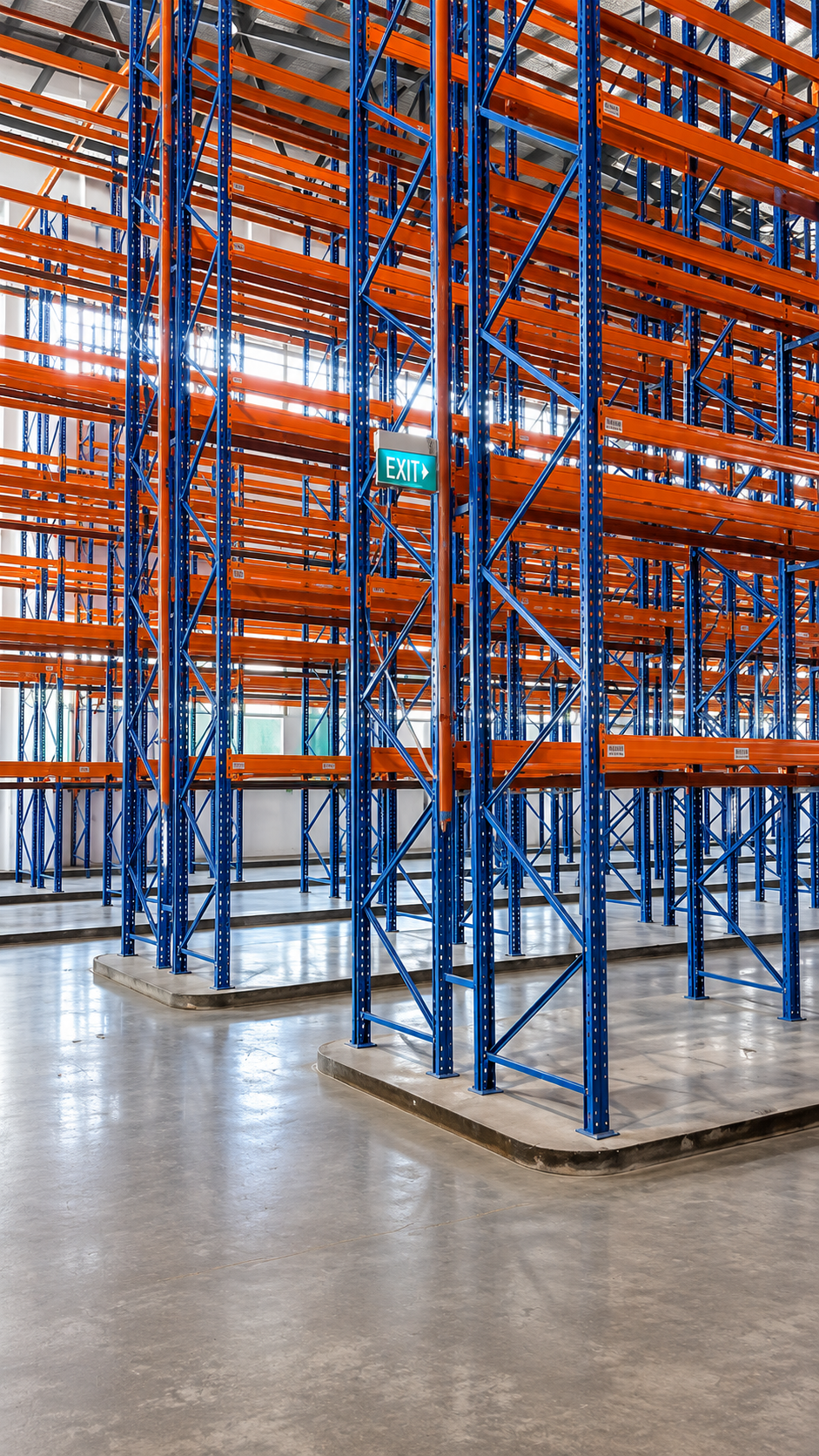 Empty warehouse shelves with blue and orange metal racks, an exit sign, and a concrete floor.