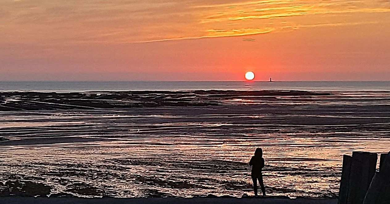 A person standing on the beach during sunset, with the ocean and a lighthouse in the distance.