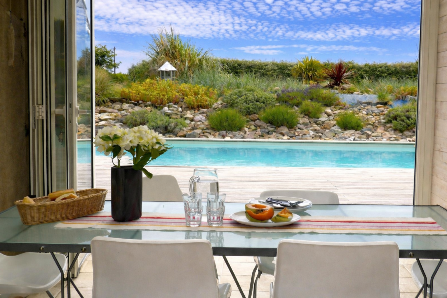A dining table set with white chairs, a vase of white flowers, a tray of bread, a bowl of sliced fruit, a pitcher of water, and glasses, with a view of a swimming pool and landscaped garden with rocks and plants through open sliding glass doors.