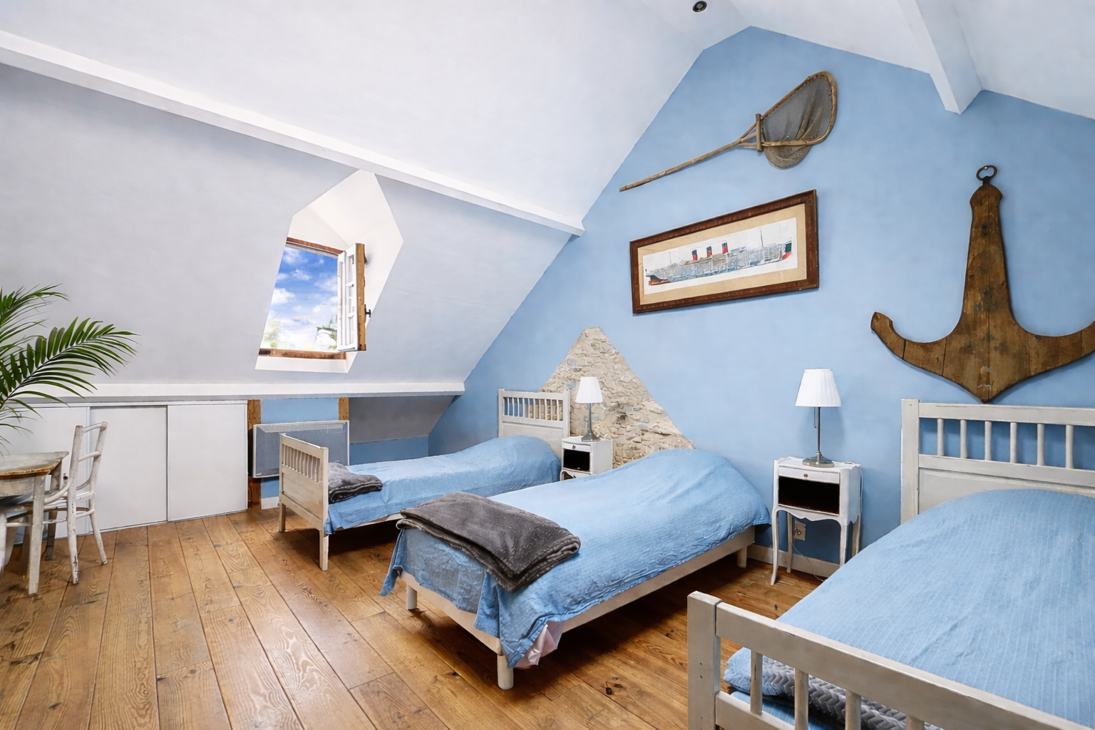 Bedroom with three single beds with light blue bedspreads, nautical decor including a large anchor, framed boat picture, and fishing net with pole. Wooden flooring, small white nightstands with lamps, window with wooden shutters letting in natural light, wooden table and chairs on the left. Spacious attic room with sloped ceiling, painted light blue and white walls, and potted plant.