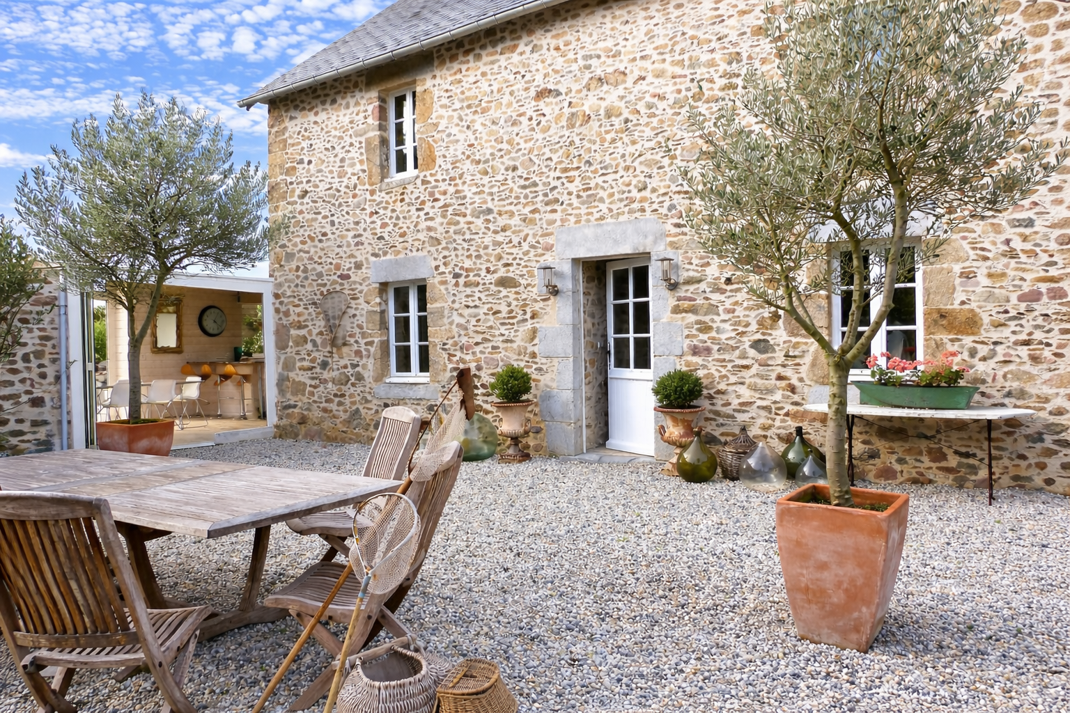 Stonehouse with a gravel patio, wooden outdoor dining table, potted trees, and decorative jars, with an open view into a bright indoor kitchen area.