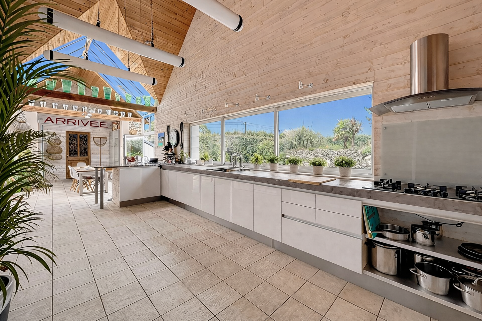 Bright kitchen with white cabinets, large window with outdoor view, wooden walls and ceiling, tiled floor, modern appliances, potted plants on the windowsill, and an 'Arrivee' sign in the background.