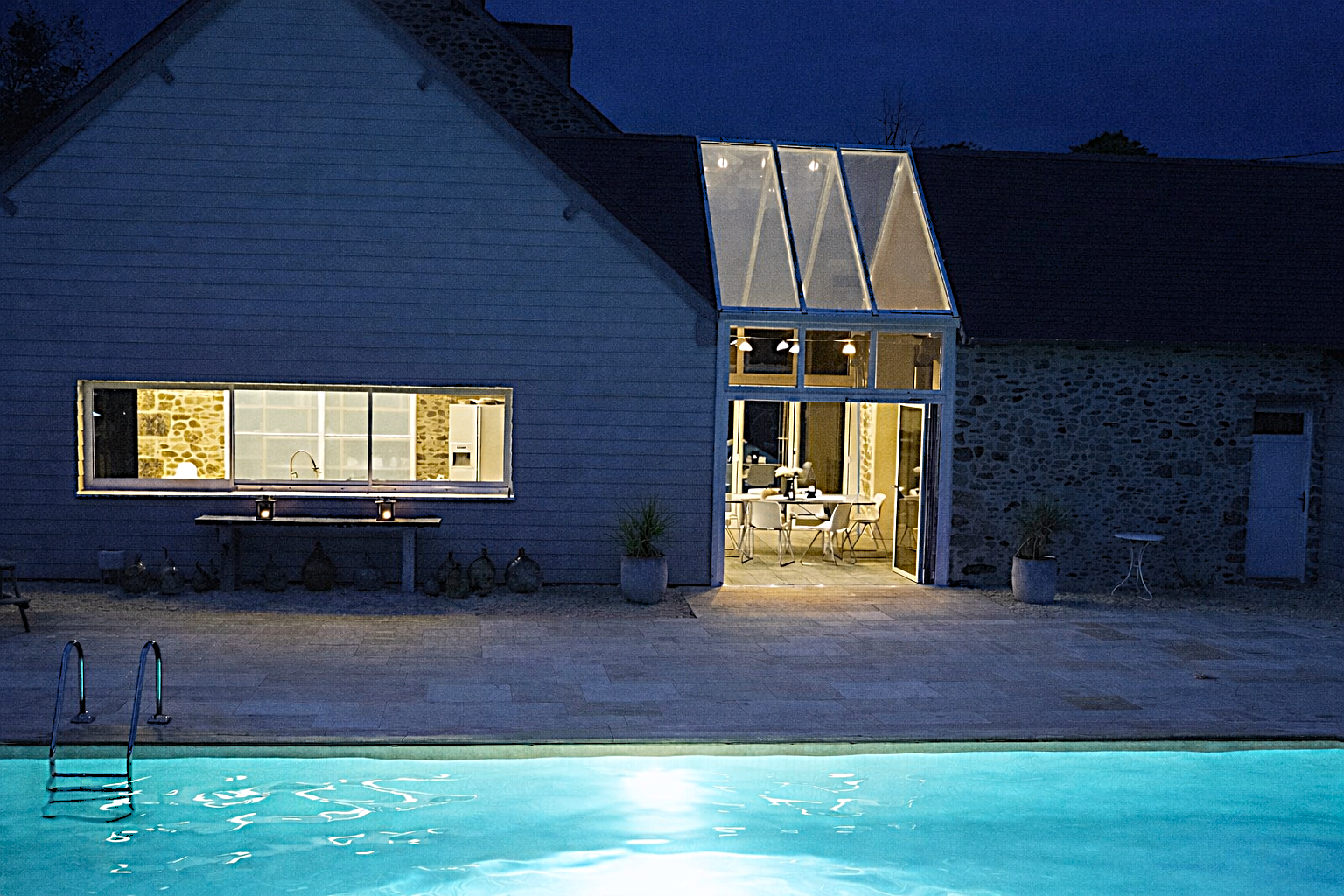 A nighttime view of a modern house with a lit interior, a large glass door opening to a patio with a swimming pool, and a glass enclosed upper section.