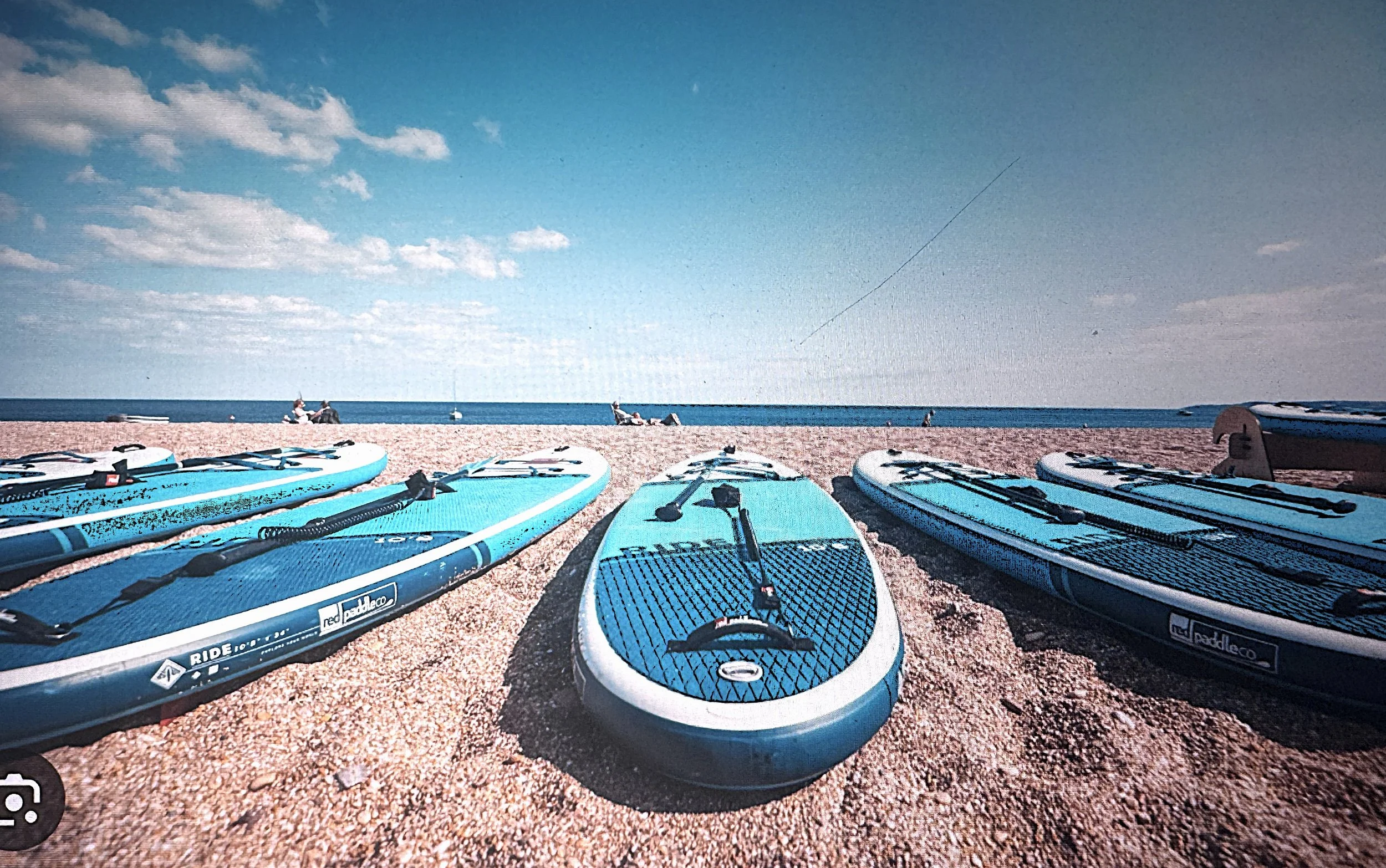 Multiple blue stand-up paddleboards lined up on a gravel beach with a few people sitting in the background, a blue sky with scattered clouds, and the ocean in the distance.