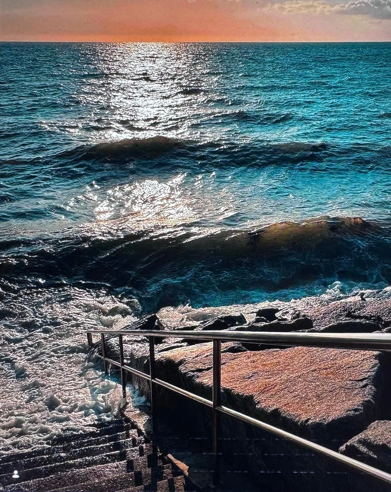 Sunset over the ocean with waves crashing against rocks and a metal railing in the foreground.