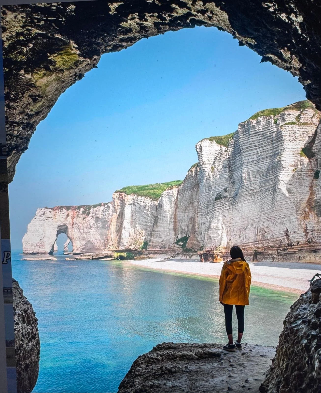A person in a yellow jacket stands on a rocky ledge inside a sea cave, looking out at white chalk cliffs and a natural arch formation along a beach under a clear blue sky.