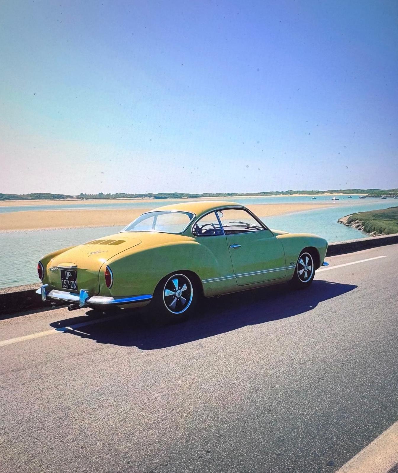 A vintage yellow-green sports car parked on a coastal road near water with small boats, sandy beach, and green landscape in the background under a clear blue sky.