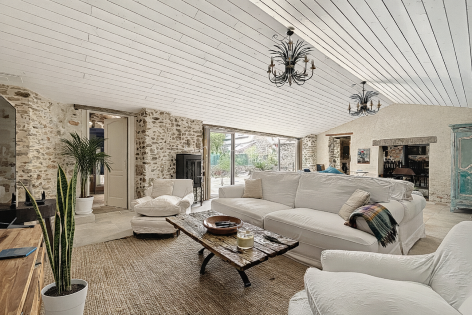 Living room with white sofas, stone walls, wooden coffee table, potted plants, and large windows showing outdoor patio.