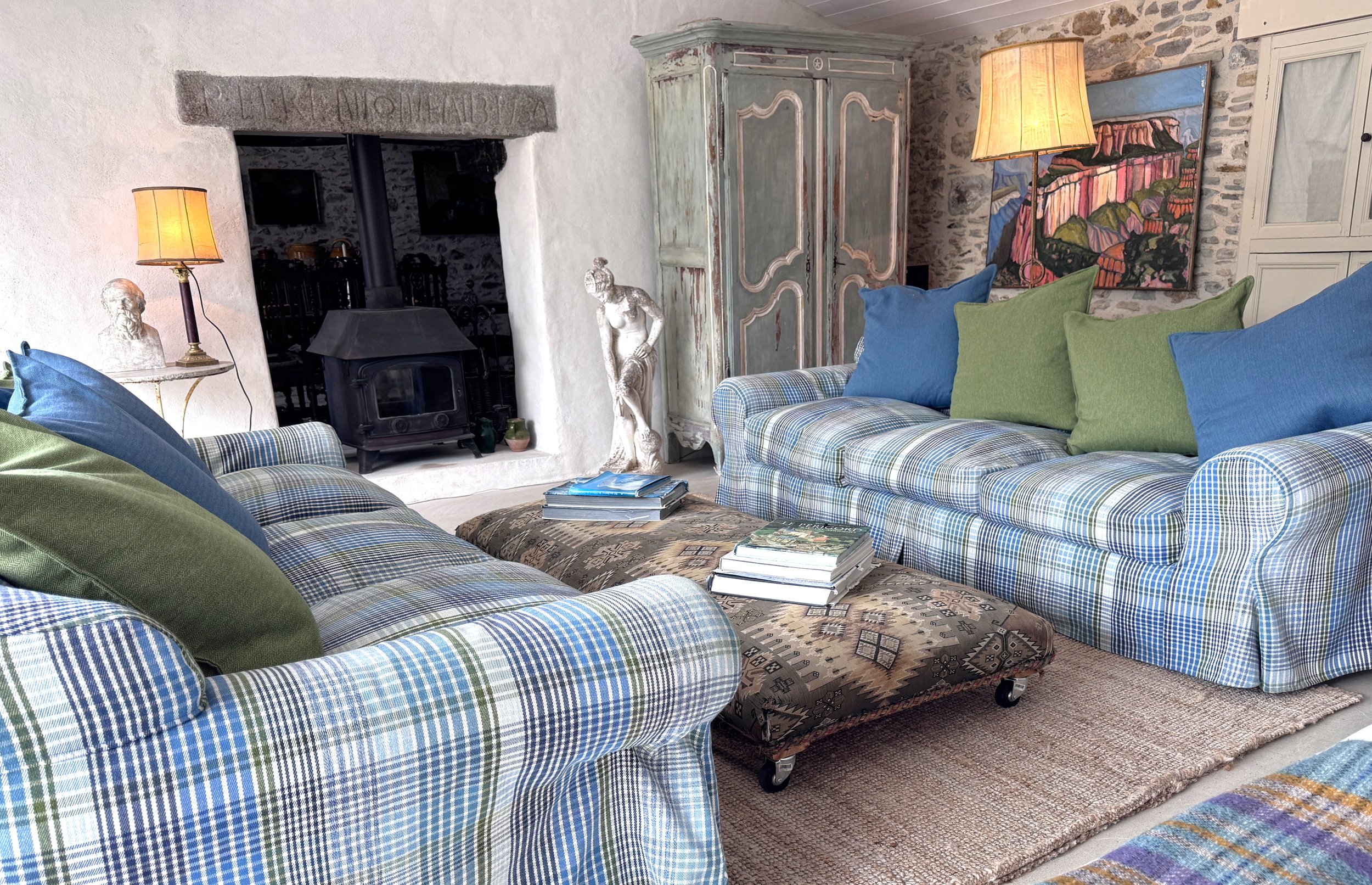Living room with stone walls, a plaid sofa with blue and green pillows, a wooden coffee table with magazines, a fireplace, a vintage wardrobe, a large abstract landscape painting, and table lamps.