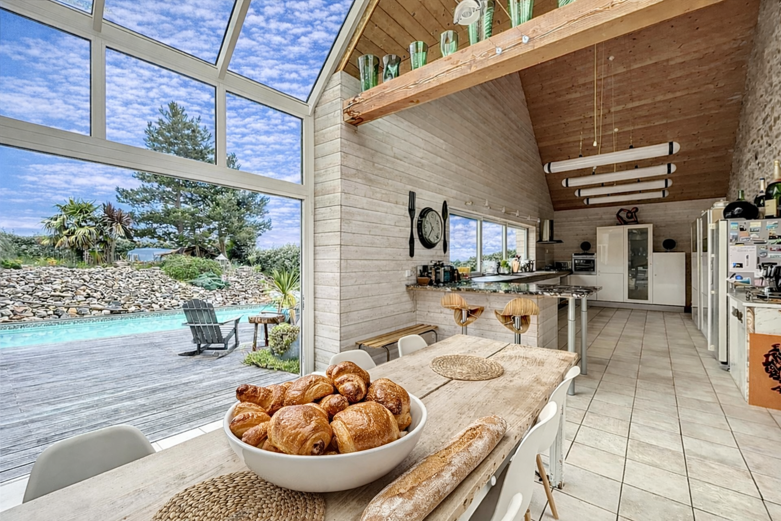 Indoor kitchen with large windows showing backyard pool and garden, wooden ceiling, and dining table with croissants