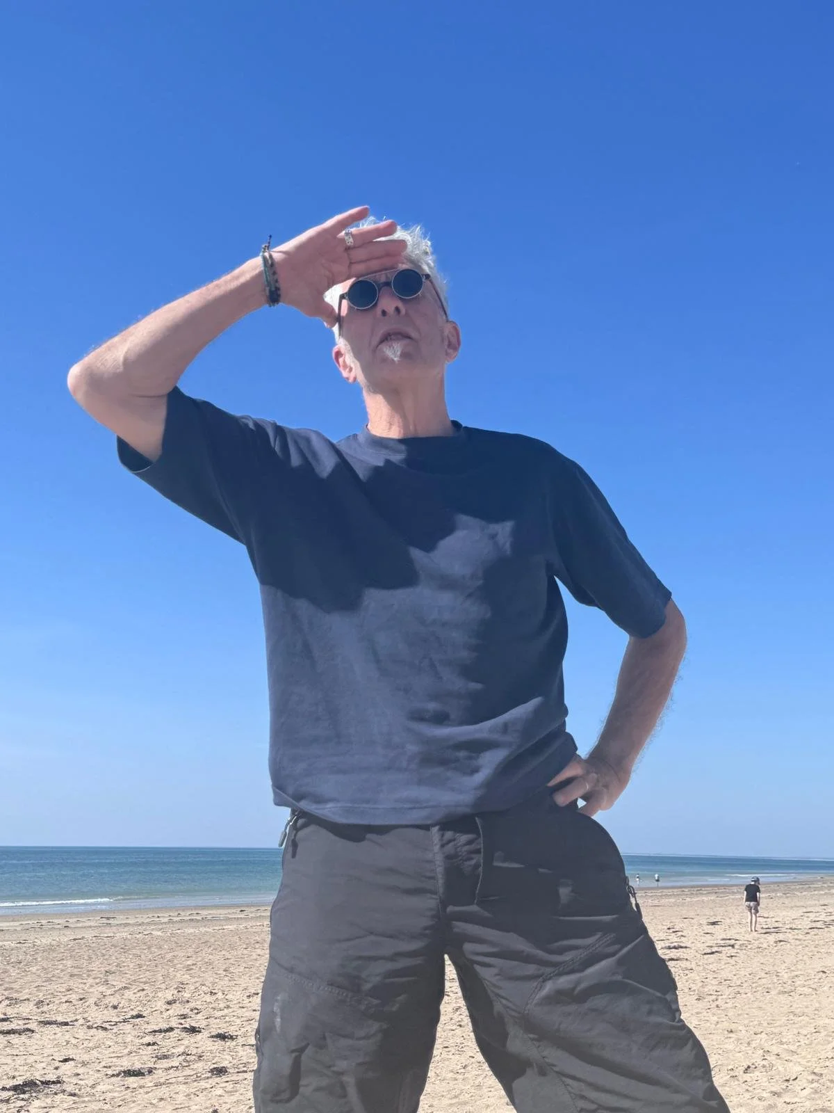 A man with gray hair and sunglasses standing on a sandy beach, shielding his eyes from the sun with his hand, with the ocean and clear blue sky in the background.