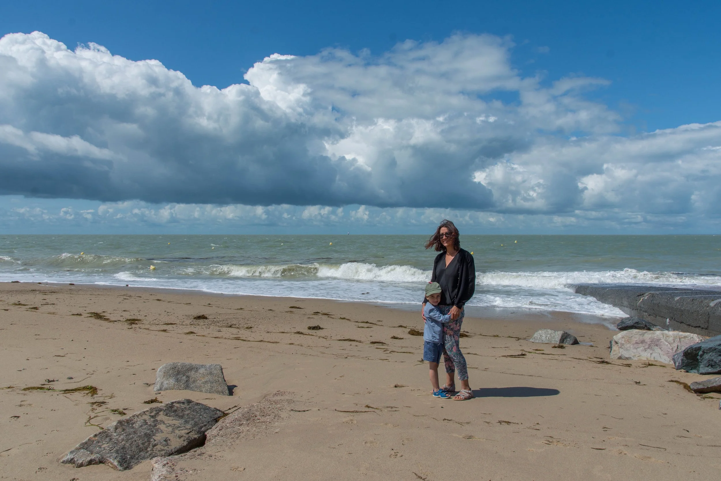 A woman and a young girl standing on a sandy beach with rocks, with the ocean and clouds in the background.