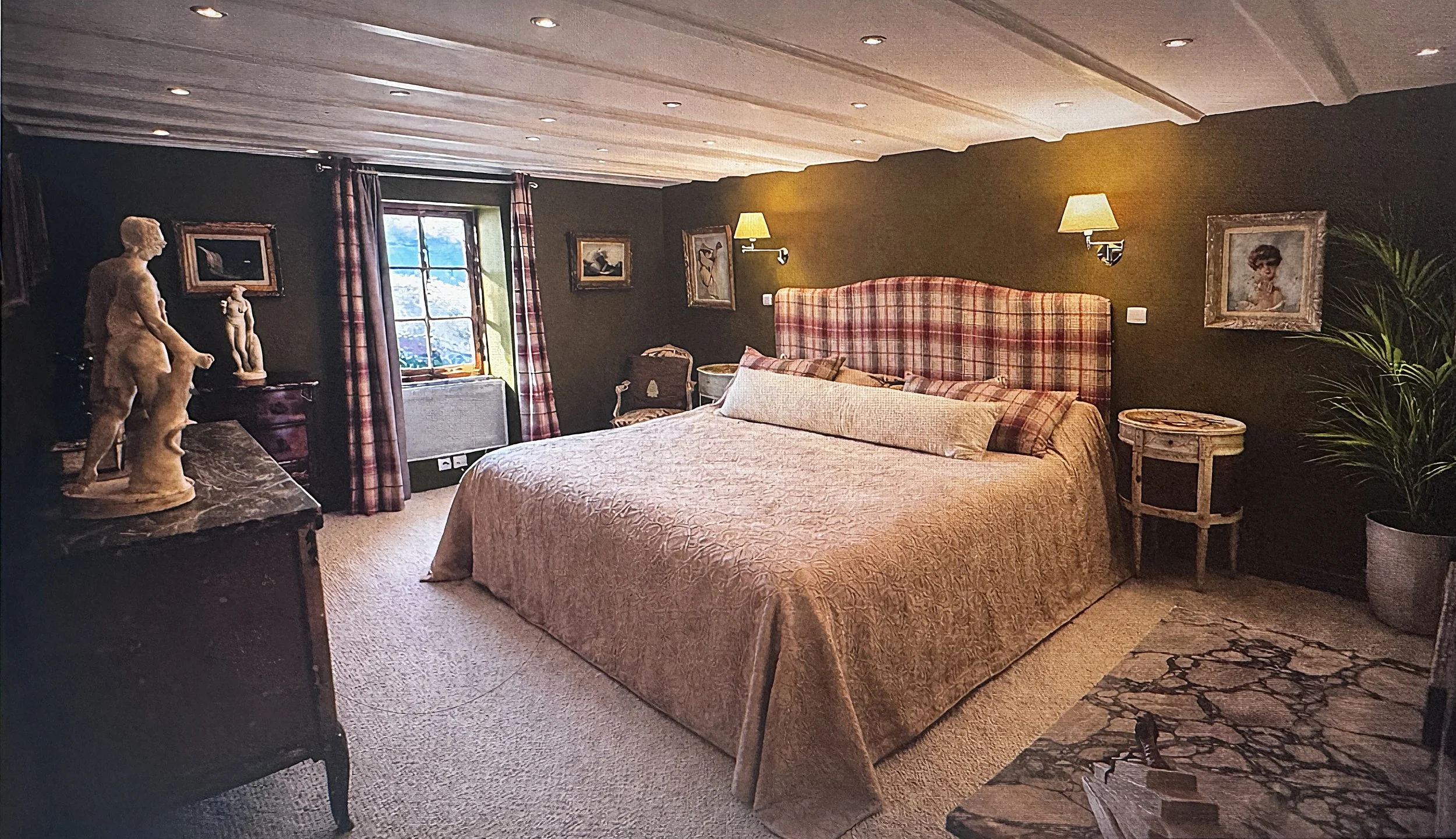 A cozy bedroom decorated with classical art and sculptures, featuring a large bed with a plaid headboard, wall-mounted lamps, and framed artwork, with window curtains and a window showing a mountain view.