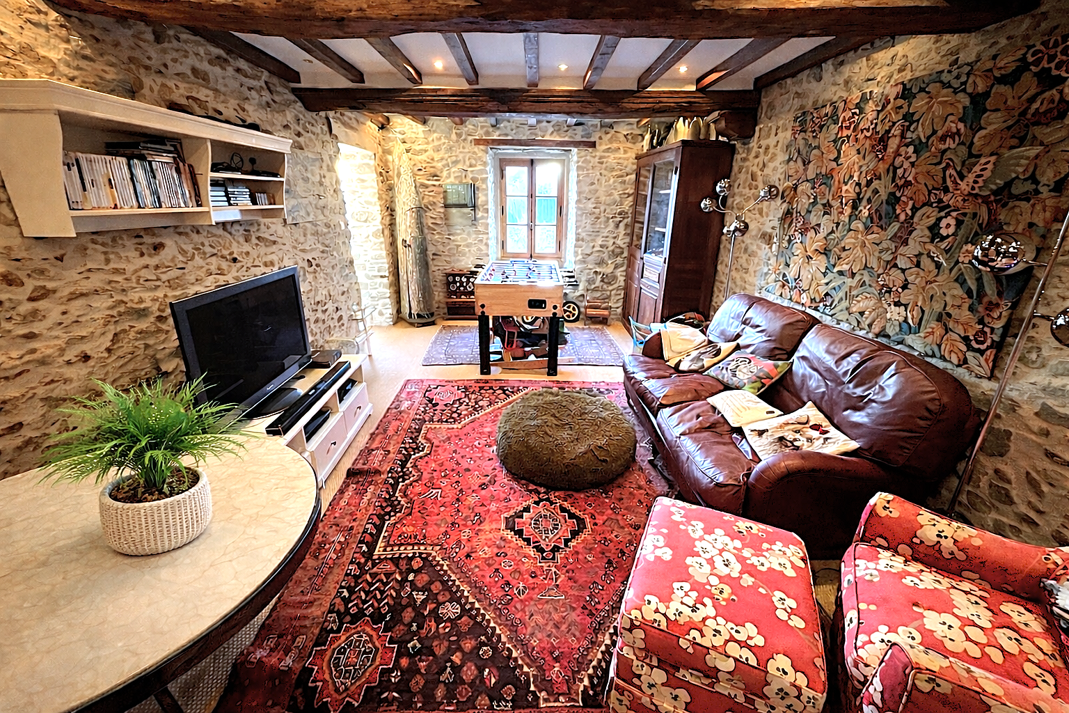 A cozy living room with stone walls, beamed ceiling, and a window that lets in natural light. The room features a brown leather sofa with pillows, a red floral armchair, a television on a white stand, a green potted plant on a table, a foosball table near the window, and various decorative items including a tapestry on the wall and books on a shelf.