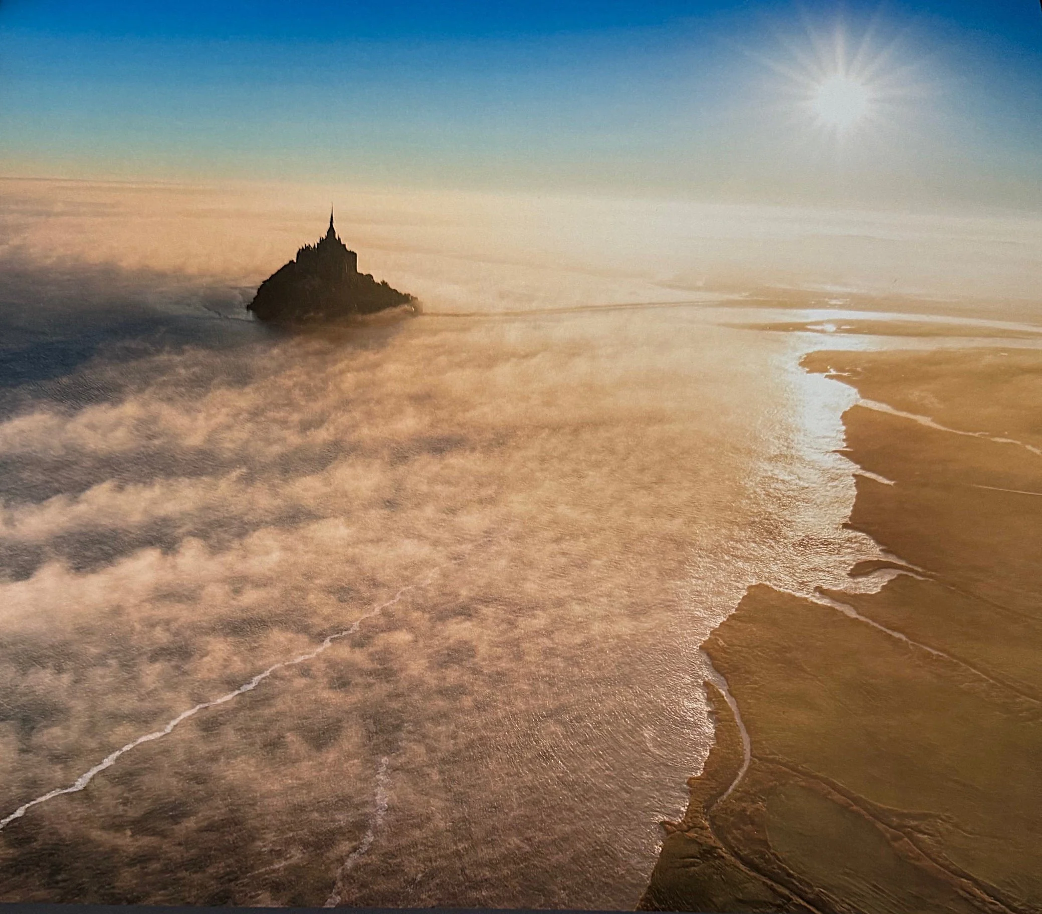 A rocky island with a spire-like structure on top, surrounded by water and clouds with a bright sun in the sky.