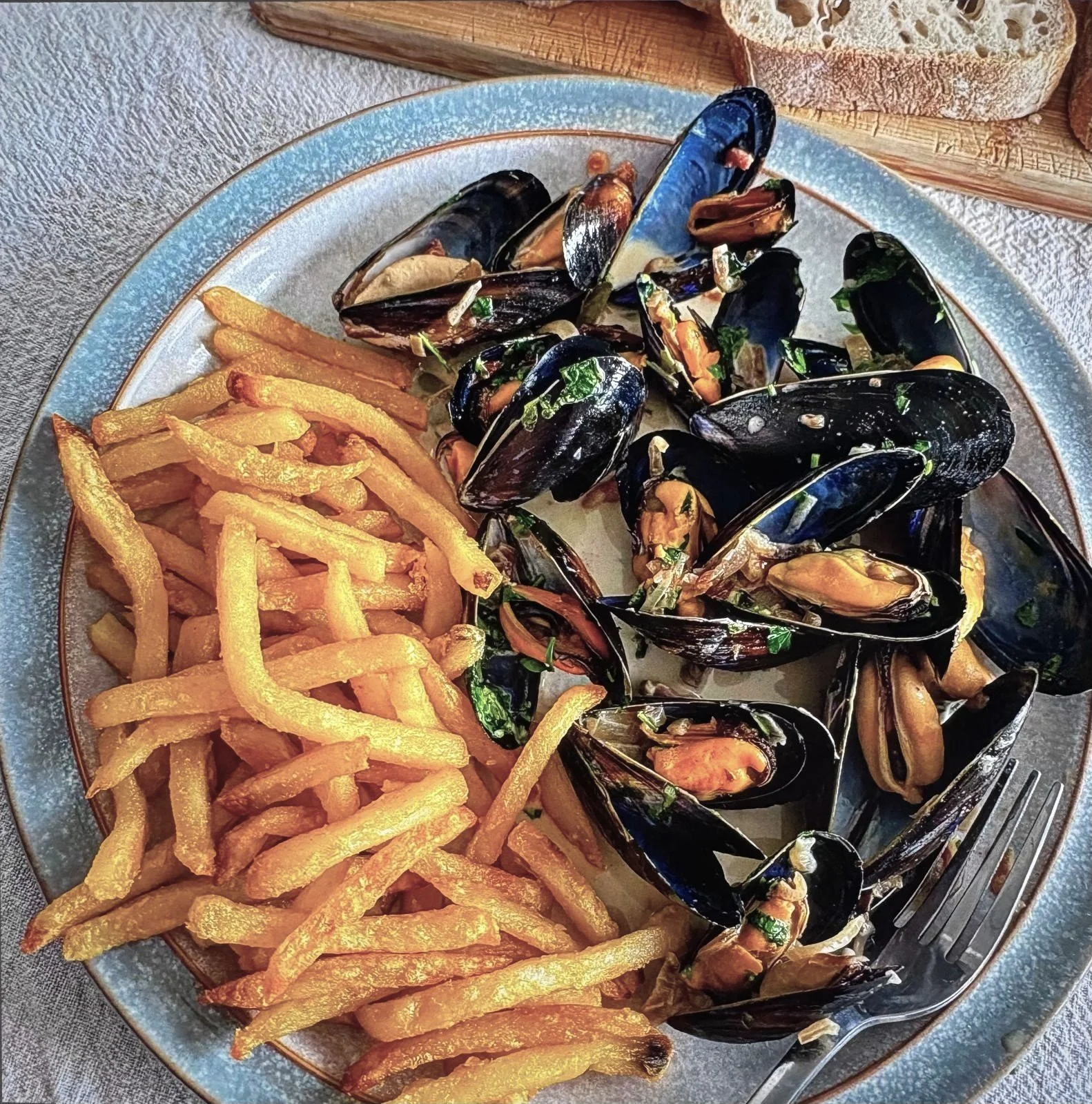 Plate of cooked mussels with herbs, served alongside French fries, on a textured table with a fork nearby.
