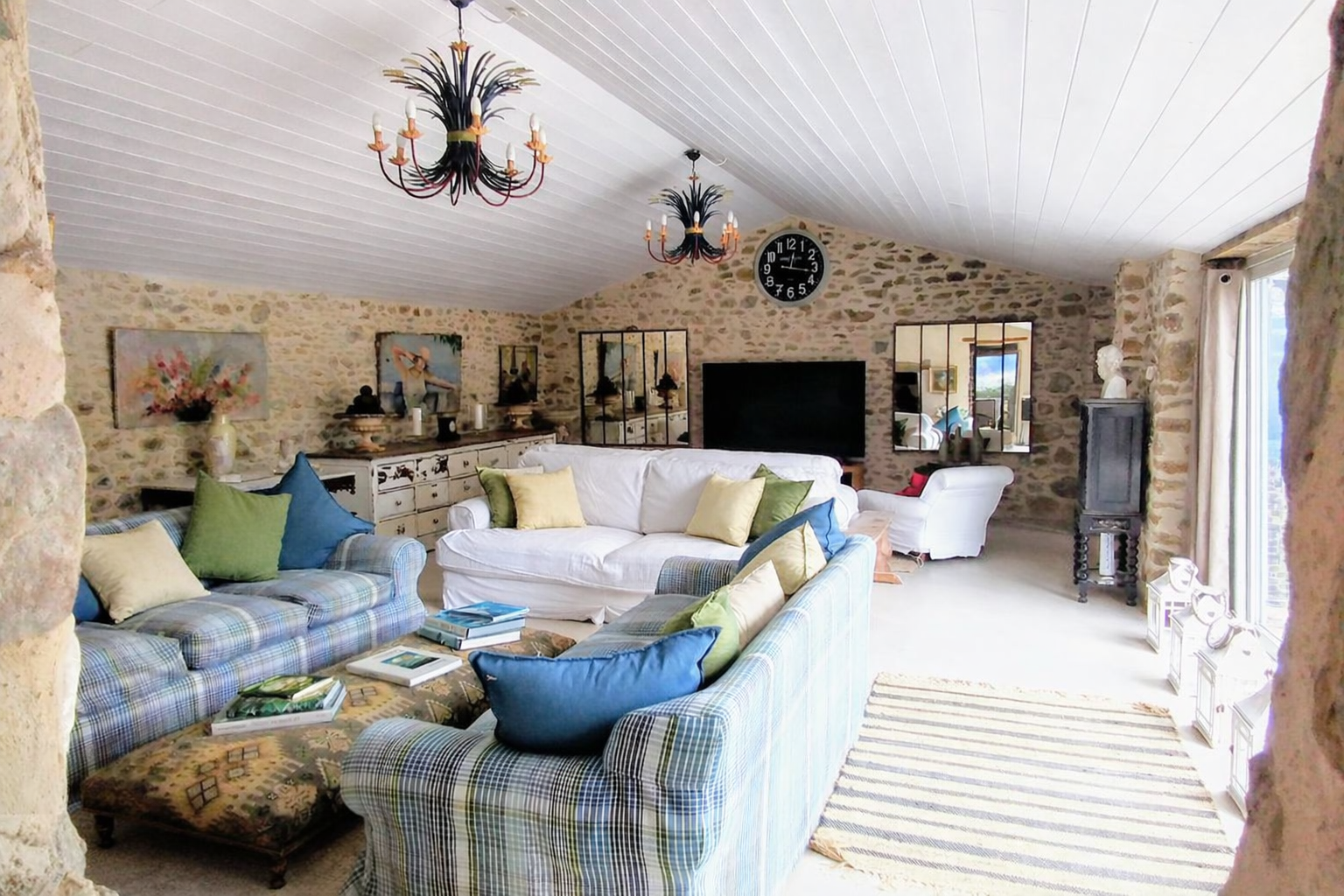 Living room with stone walls, white ceiling, and large window with curtains. Contains colorful sofas with blue, green, and beige cushions, a coffee table with books, a TV, and decorative items. Two chandeliers hang from the ceiling, and there is a large clock on the wall.