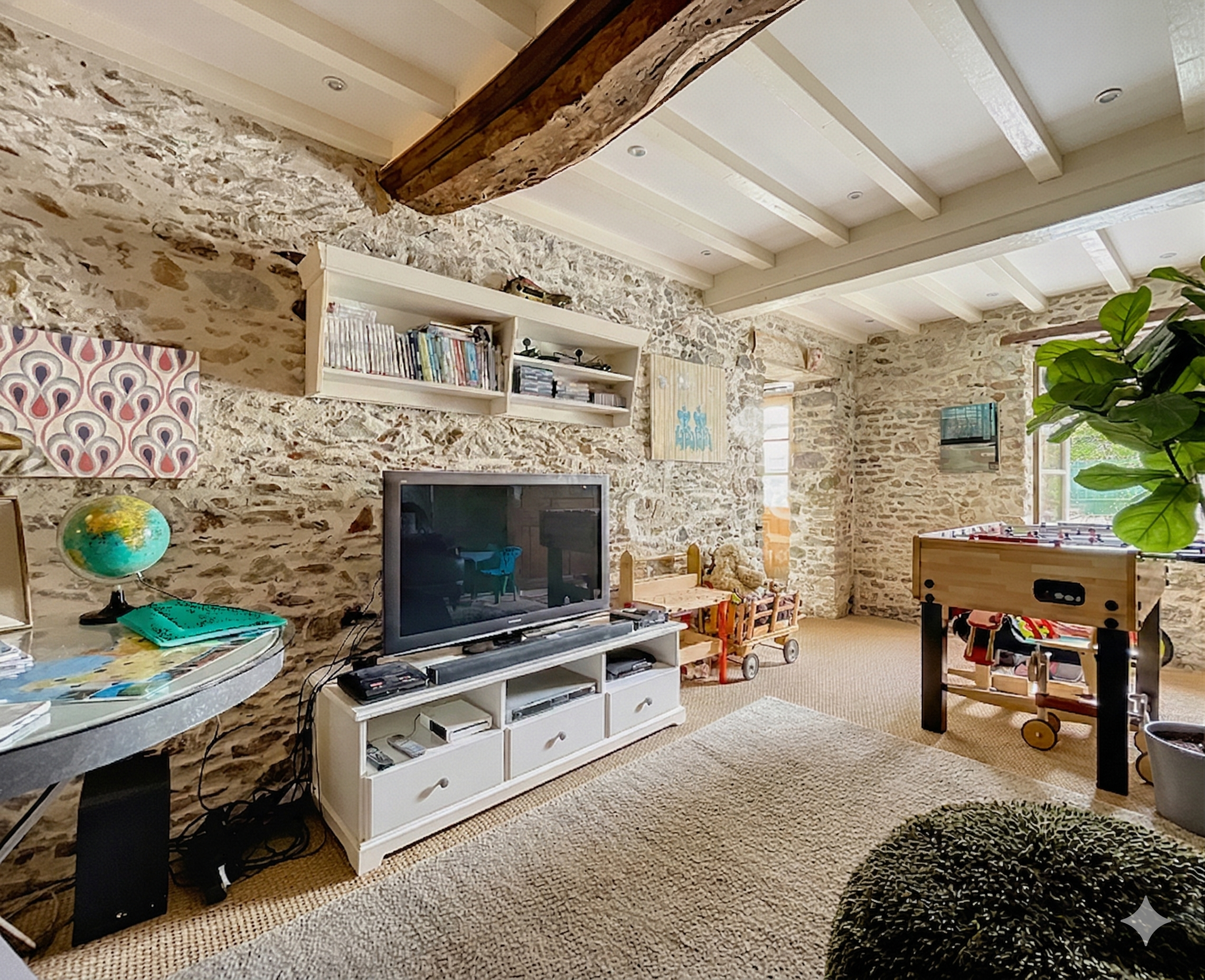 Interior of a living room with stone walls, a white bookshelf with books, a TV, a globe, a foosball table, and large windows with natural light. There is a large green plant in the corner and a beige rug on the floor.