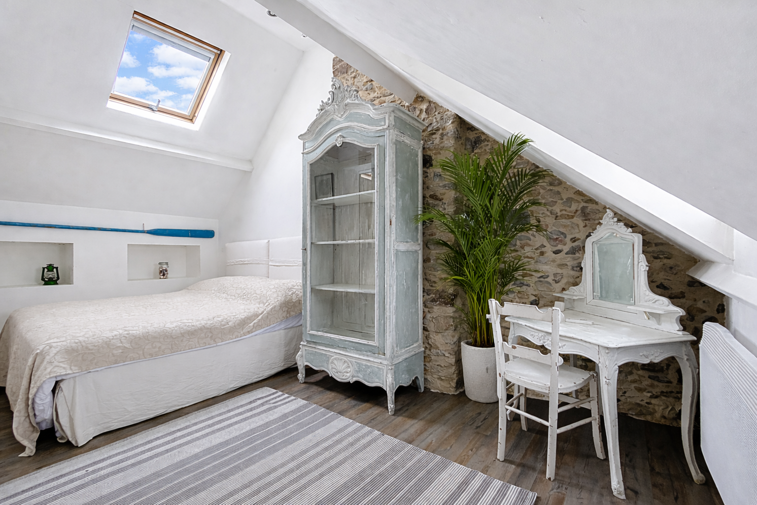 A cozy attic bedroom with a sloped ceiling, featuring a bed, vintage furniture, a potted plant, and a skylight showing a blue sky with clouds.