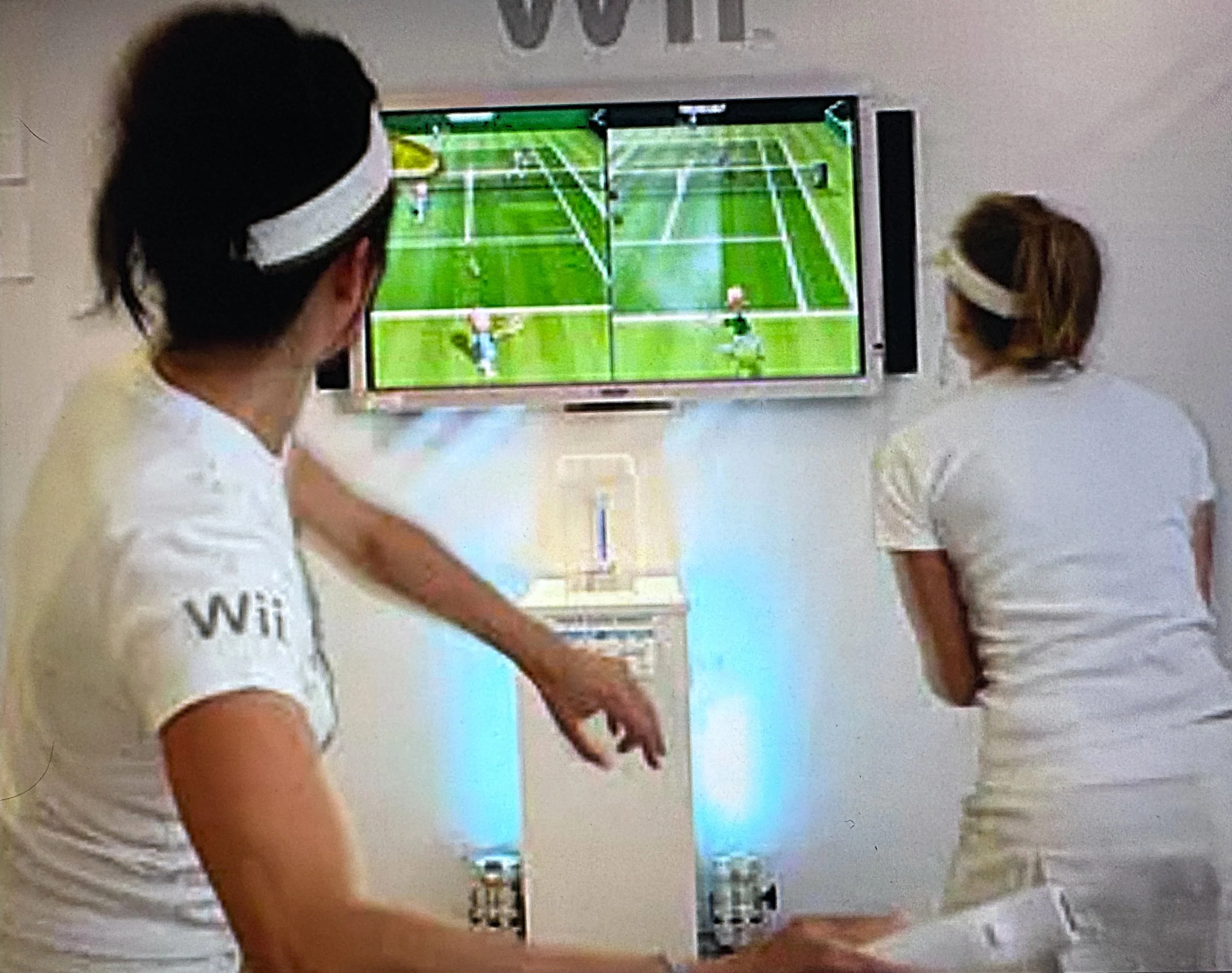 Children in white t-shirts playing tennis on indoor courts while watching a TV showing a tennis match.
