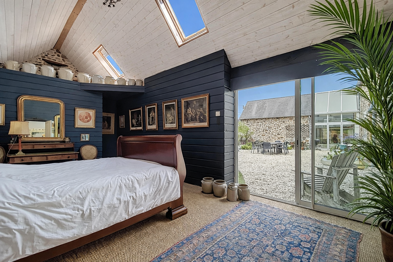 A bedroom with a wooden bed frame, white bedding, a blue accent wall with framed artwork, a dresser with a gold-framed mirror, a lamp, and a vase. Large sliding glass doors open to a gravel outdoor patio with seating. High ceiling with skylights and exposed beams, with potted plants and an area rug.