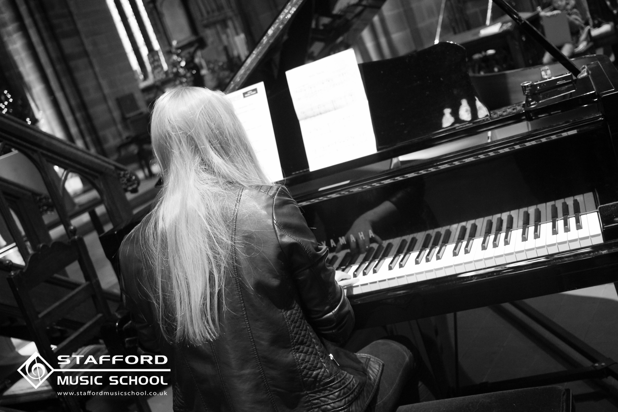 A woman with long light-colored hair sitting at a black grand piano with the lid open, playing music in a music school. The image is in black and white with the Stafford Music School logo in the bottom left corner.