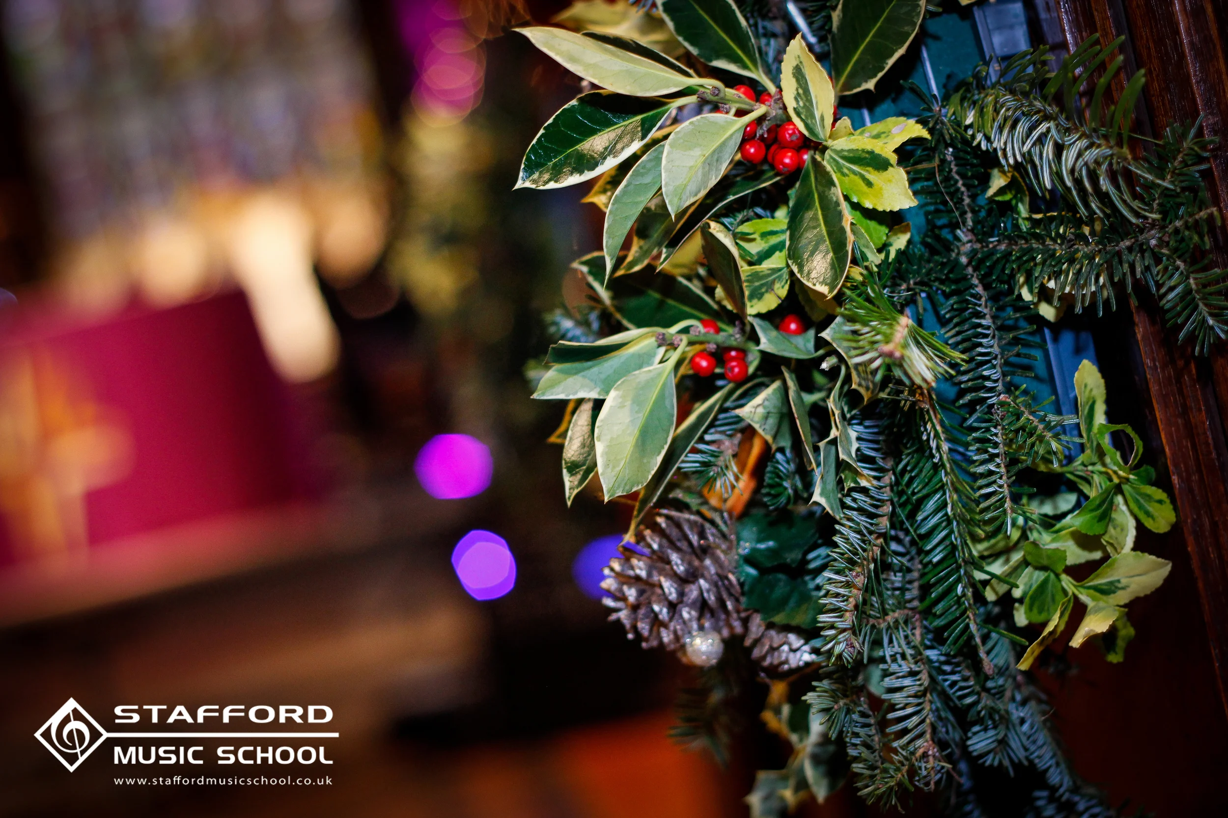 A Christmas decoration featuring holly leaves, red berries, pinecones, and evergreen branches, with a blurred colorful background and a Stafford Music School logo and website at the bottom left corner.