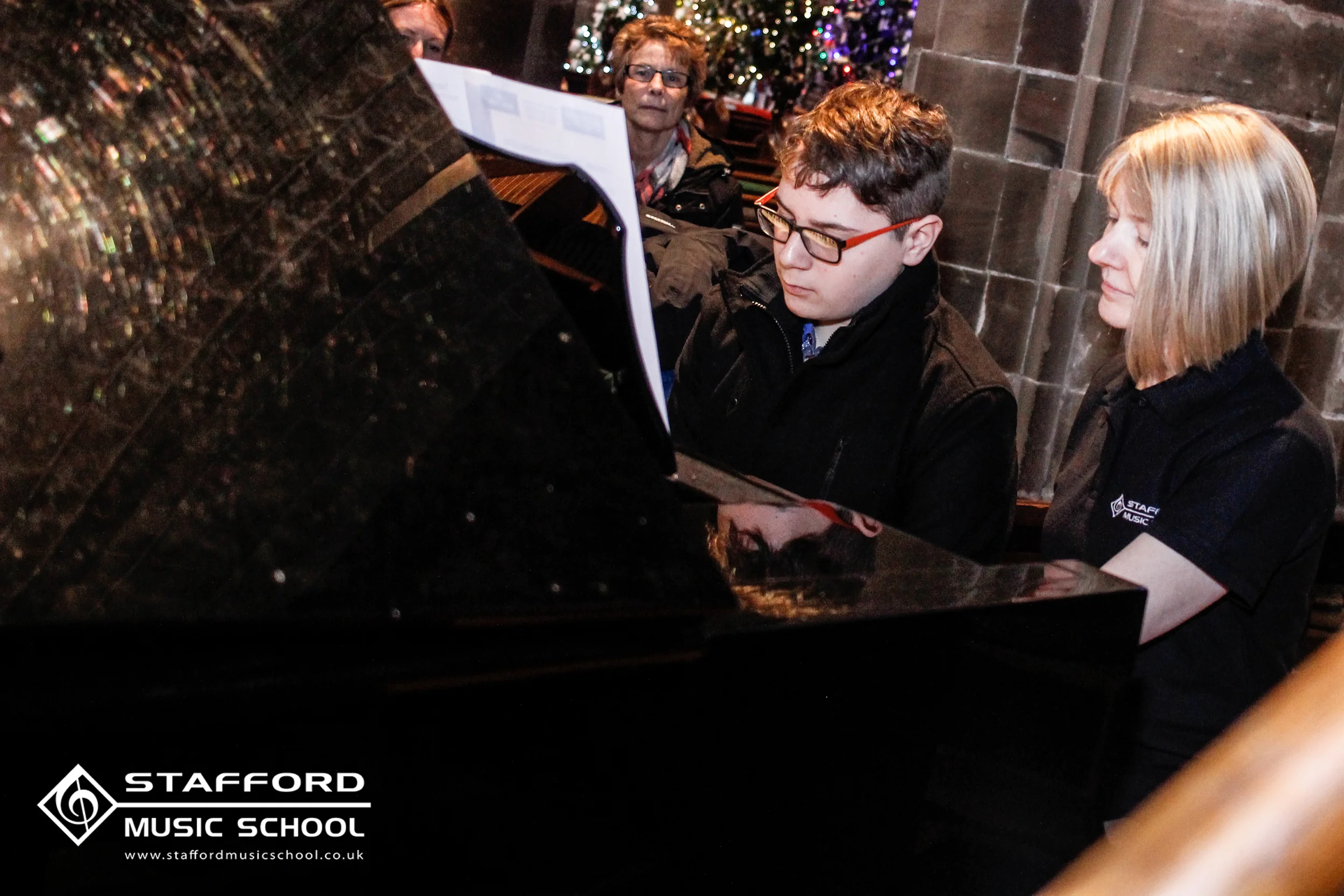 People sitting around a piano inside a decorated building with Christmas lights, at Stafford Music School.