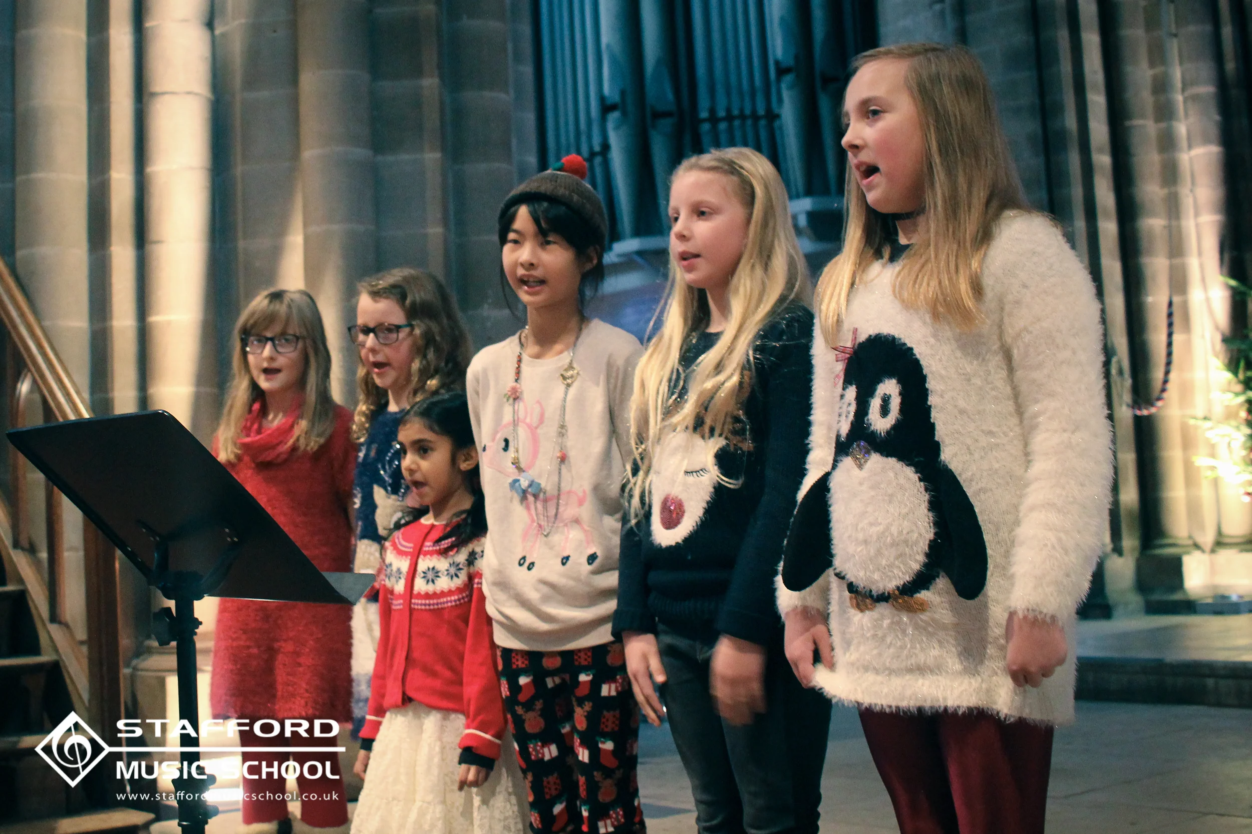 A group of young girls singing during a Christmas performance at Stafford Music School, decorated with festive holiday clothing and background with architectural columns.