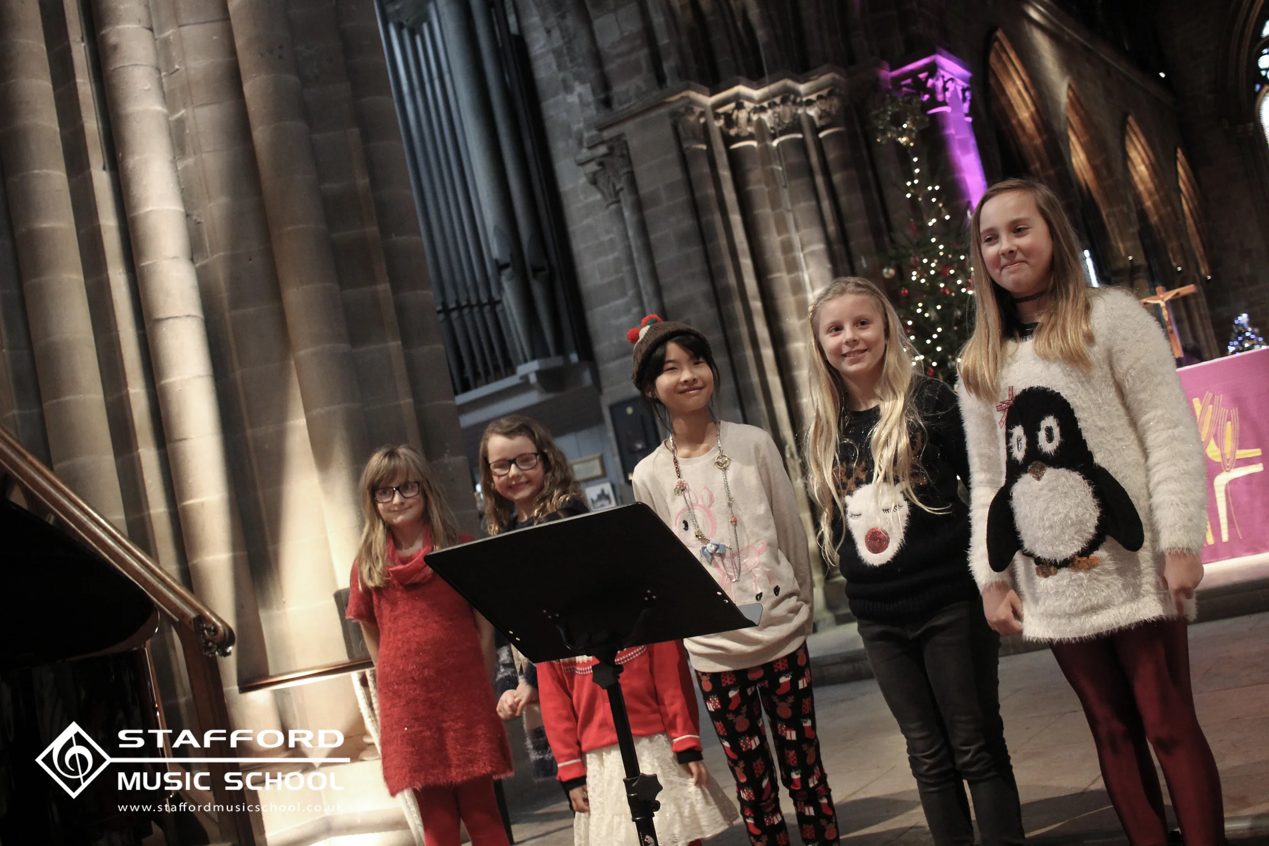 A group of young girls singing Christmas carols in a church during the holiday season, with a decorated Christmas tree in the background, and a music stand in front of them.
