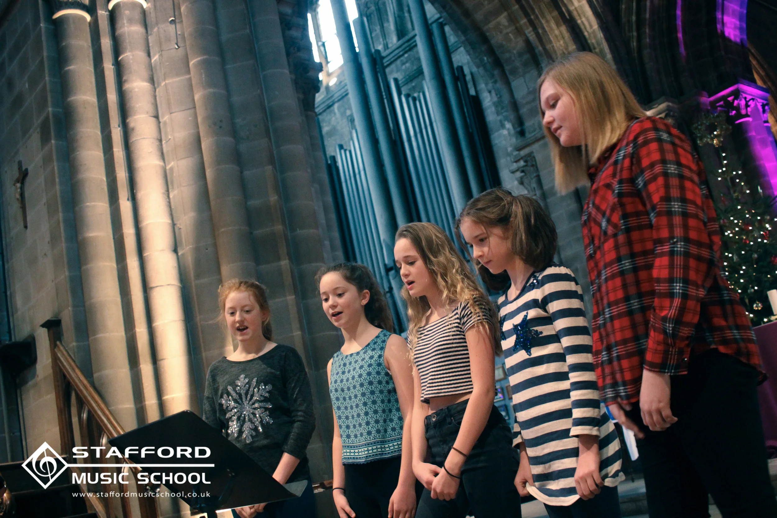 Group of children singing in a church with stained glass windows, stone columns, and purple lighting, celebrating a performance at Stafford Music School.