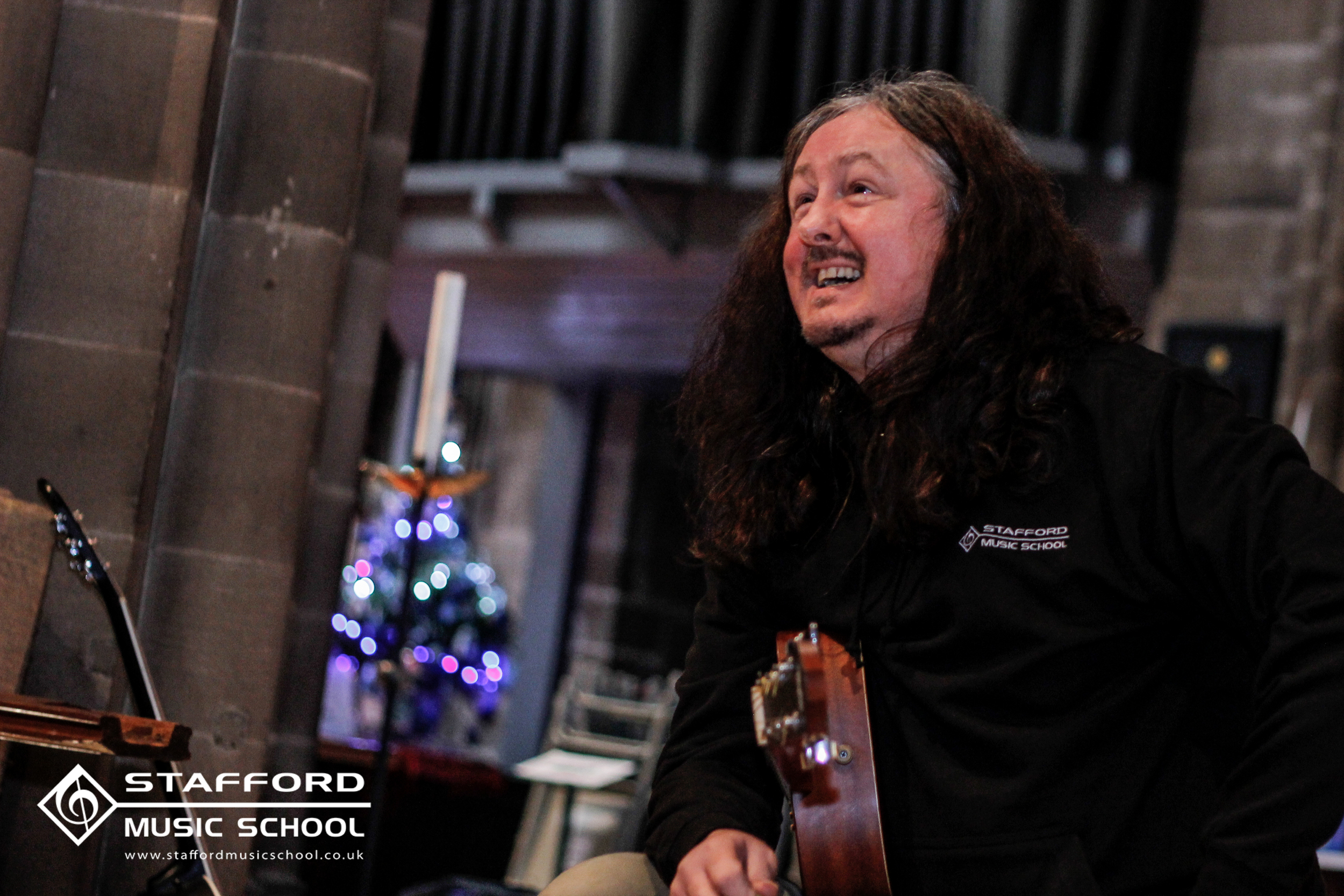 A man with long dark hair and a beard playing a guitar, wearing a black shirt with 'Stafford Music School' logo, in a room with a Christmas tree in the background decorated with lights.