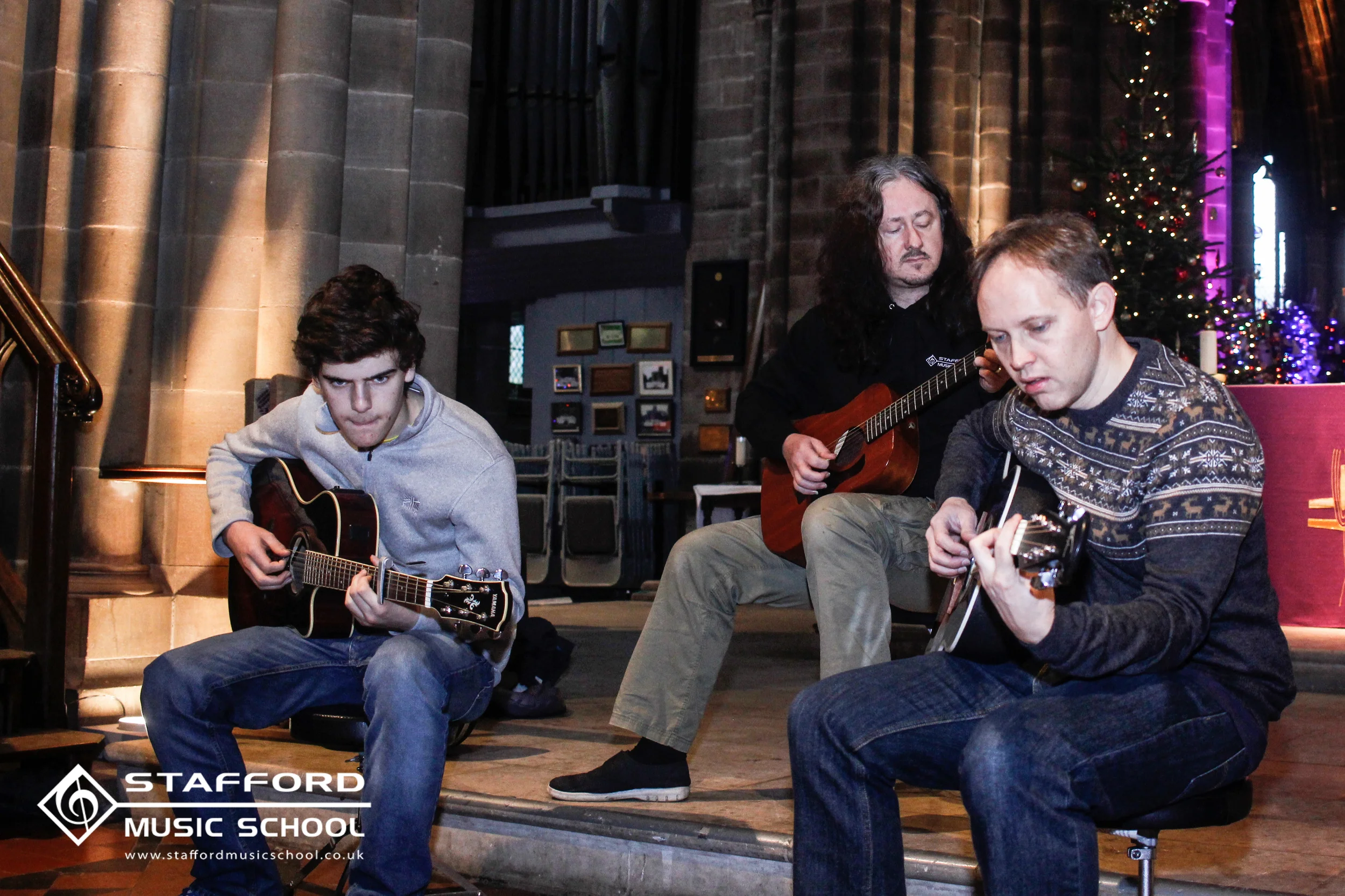 Three young men sit on a stage playing acoustic guitars. Behind them, a decorated Christmas tree and a stained glass window are visible. The scene appears to be inside a church or similar setting.