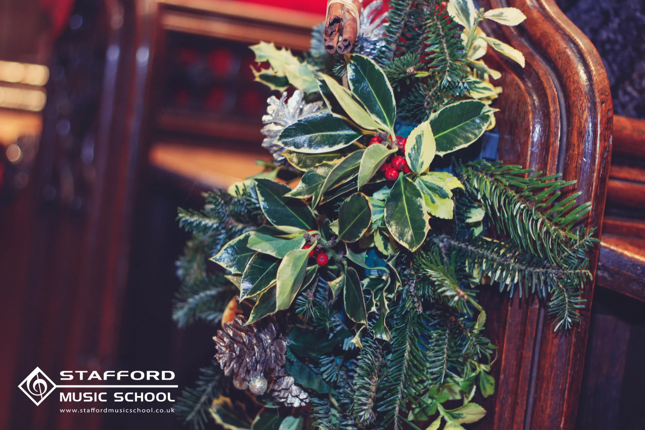 Decorative arrangement of green holly leaves, red berries, pinecones, and evergreen branches on a dark wooden surface with a logo for Stafford Music School.