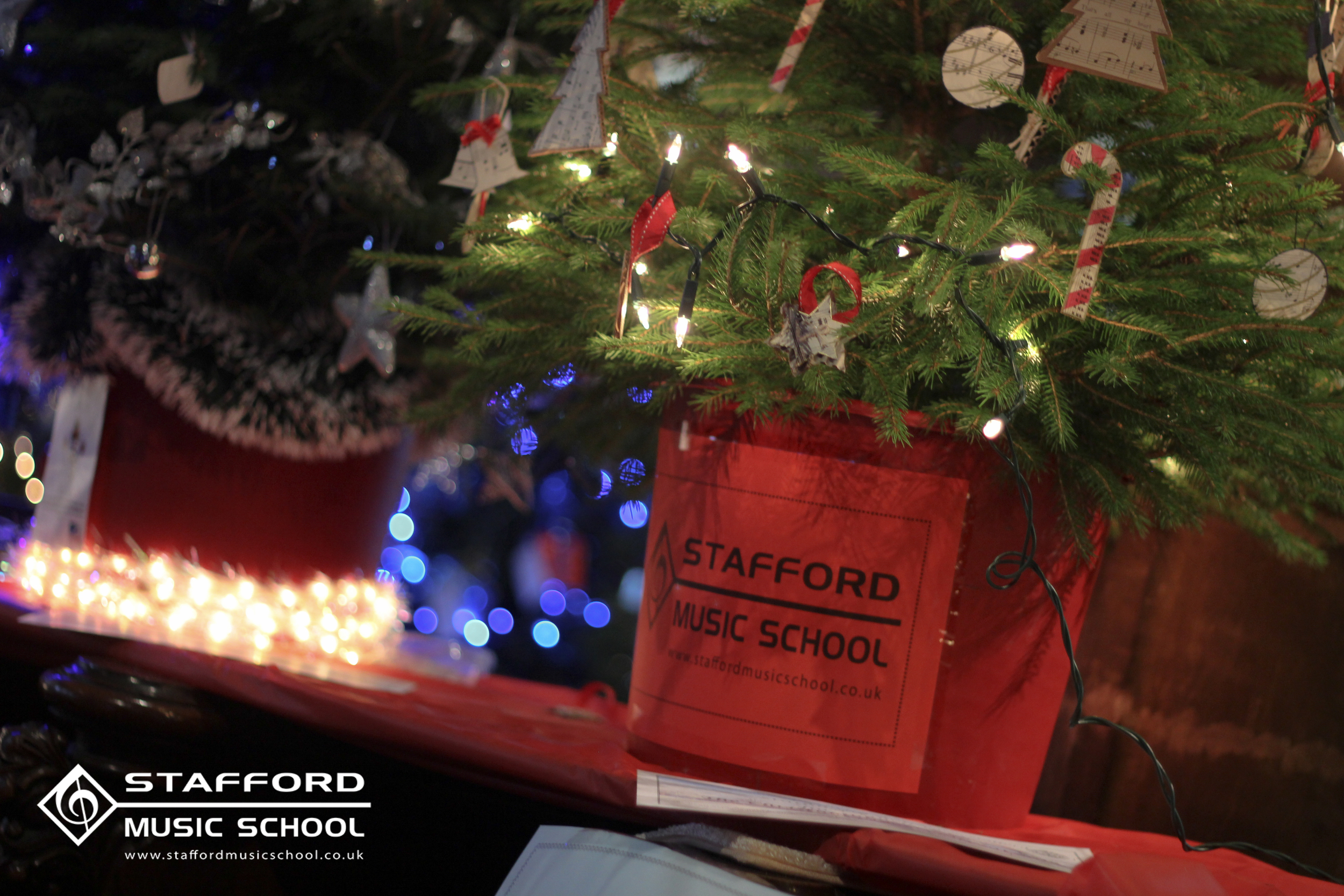 A decorated Christmas tree with ornaments and lights, in a red pot labeled 'Stafford Music School.'