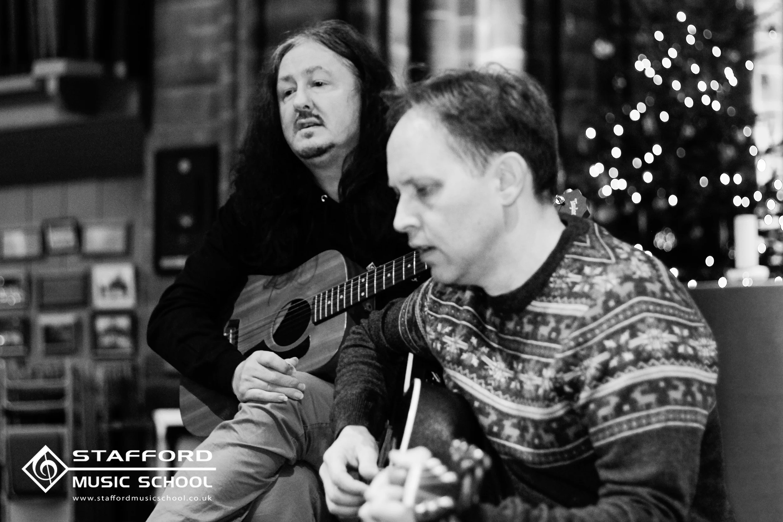 Two men playing guitars in a music classroom decorated for Christmas, with a Christmas tree in the background.
