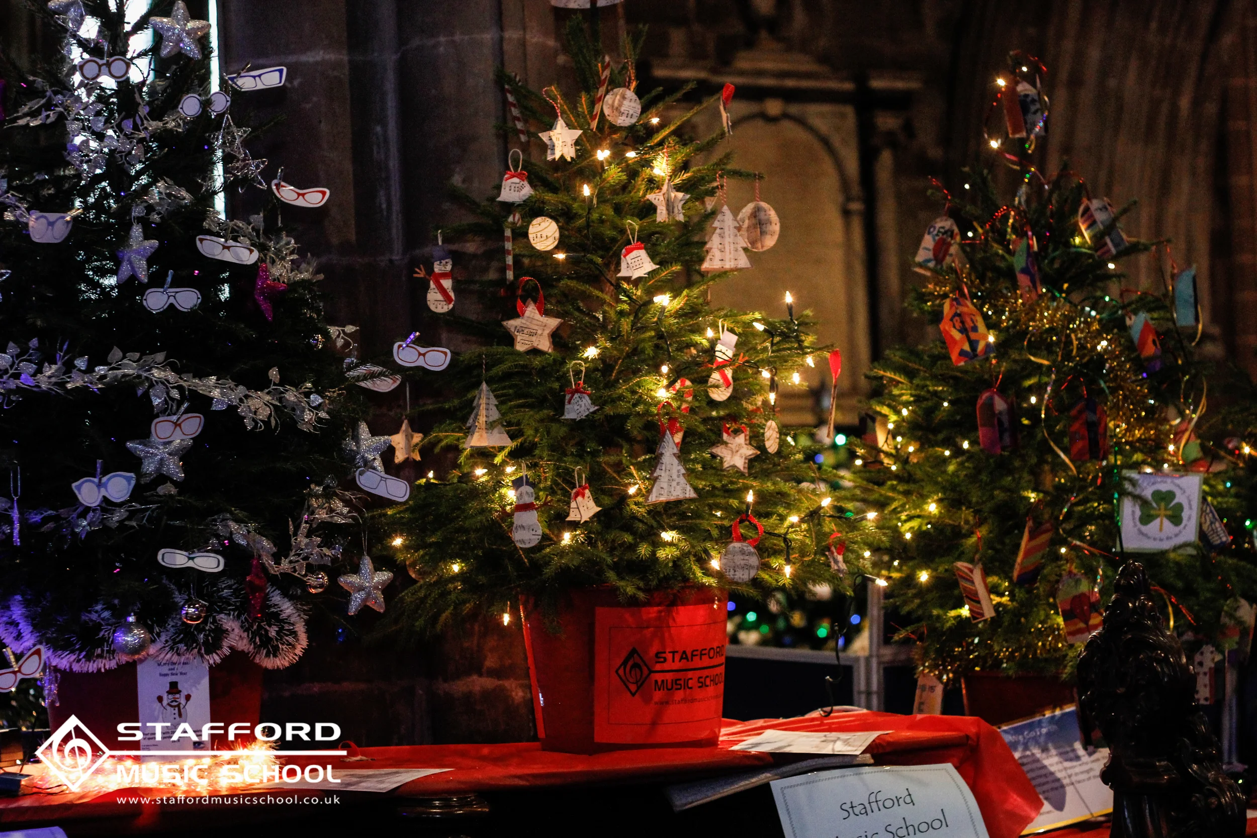 Three decorated Christmas trees with ornaments and lights, with a red Stafford Music School bucket and items on a table in front.