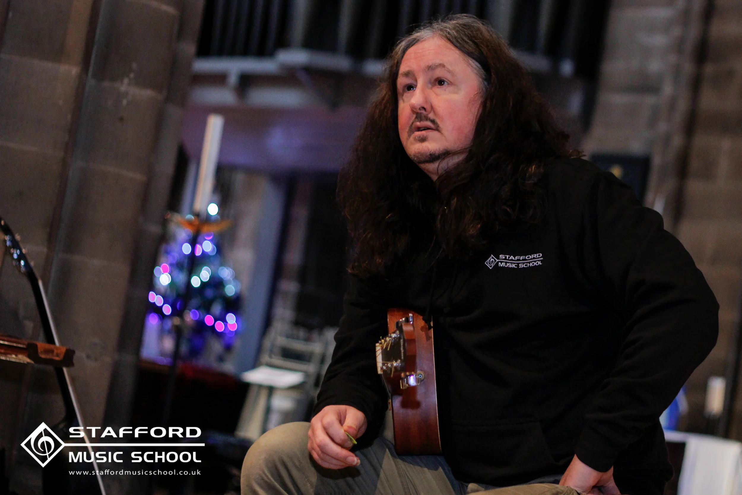 A man with long dark hair and a beard playing a guitar at Stafford Music School, with a Christmas tree in the background and holiday lights.