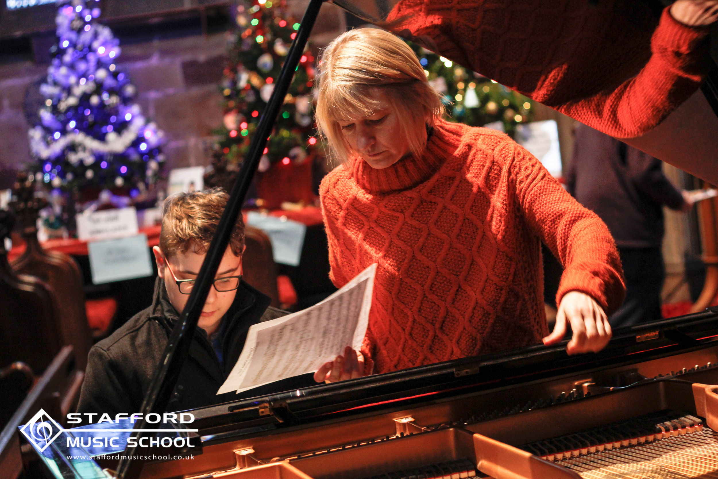 A woman with blonde hair in a red sweater standing beside a young boy with glasses, playing a grand piano during a Christmas event, with decorated Christmas trees in the background.