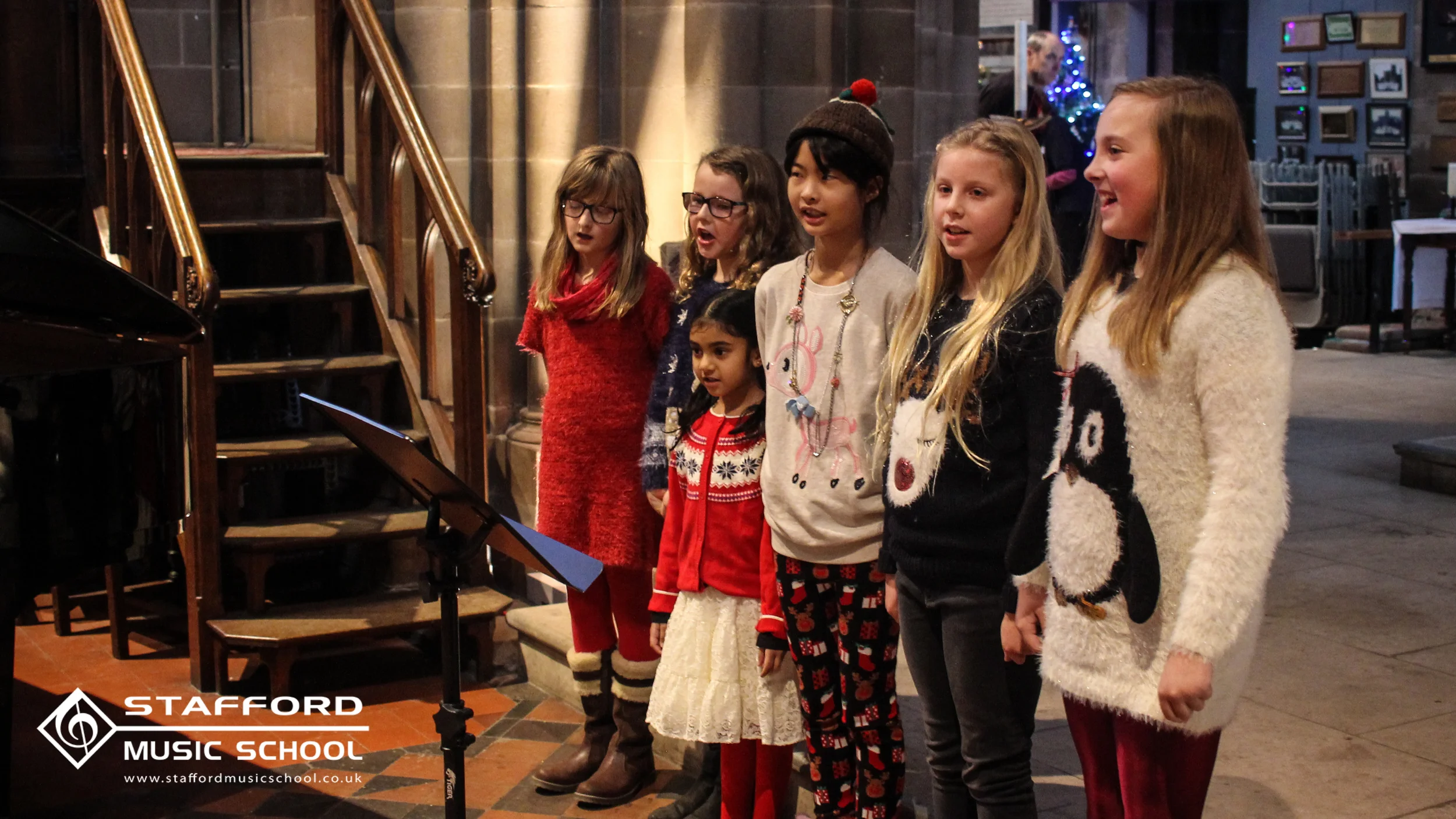 Six children singing at Stafford Music School during Christmas, with Christmas ornaments and a Christmas tree in the background.