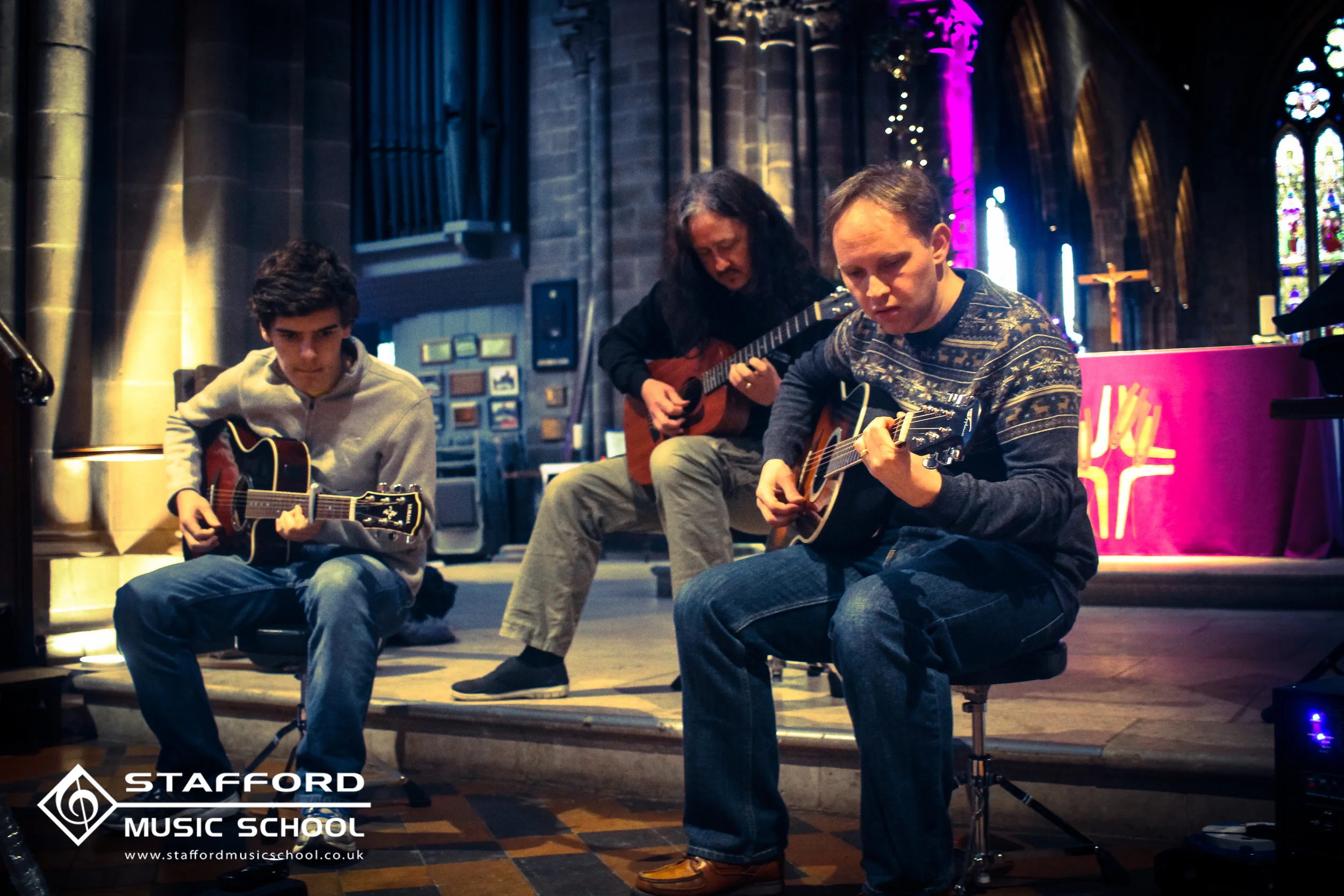 Three males playing acoustic guitars in a church setting during music rehearsal at Stafford Music School.