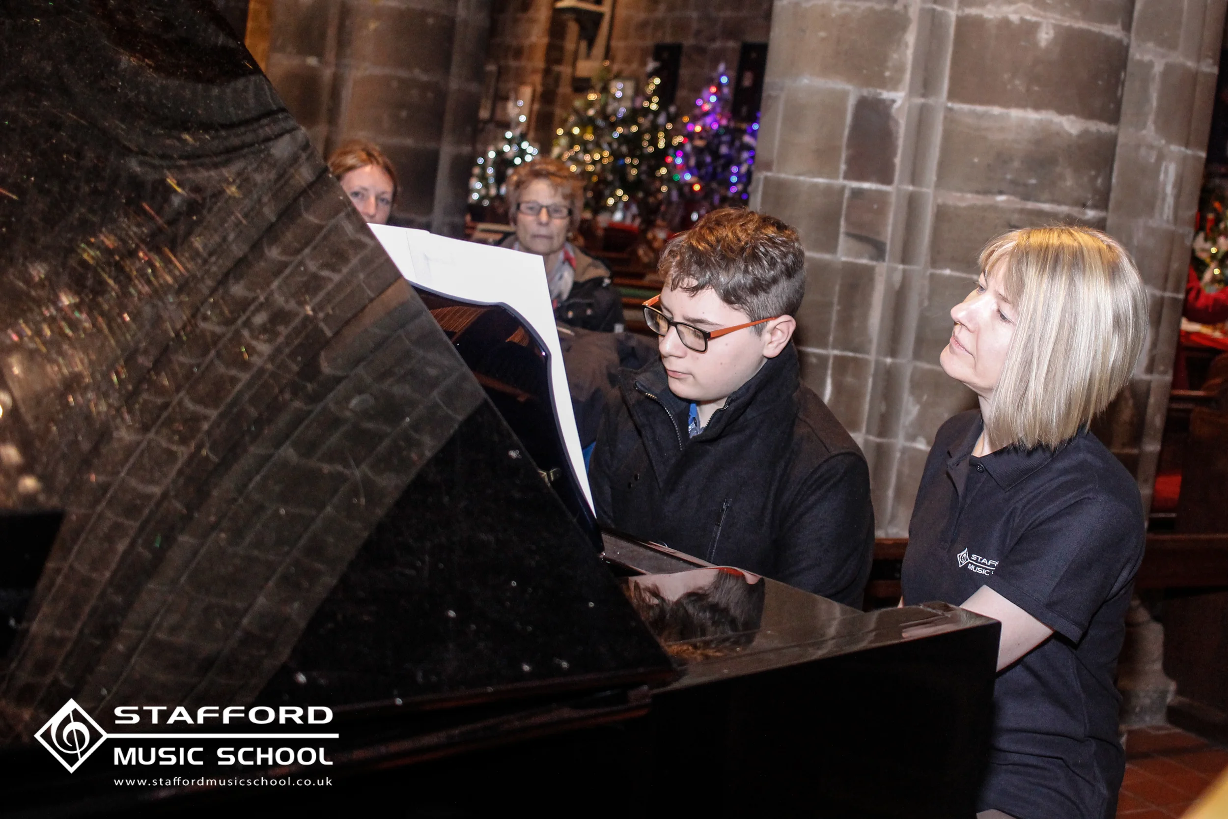 People sitting at a black grand piano with sheet music, in a room decorated for Christmas with a Christmas tree with lights and ornaments in the background. One woman and two men are visible, all wearing black shirts with "STAFFORD MUSIC SCHOOL" logo